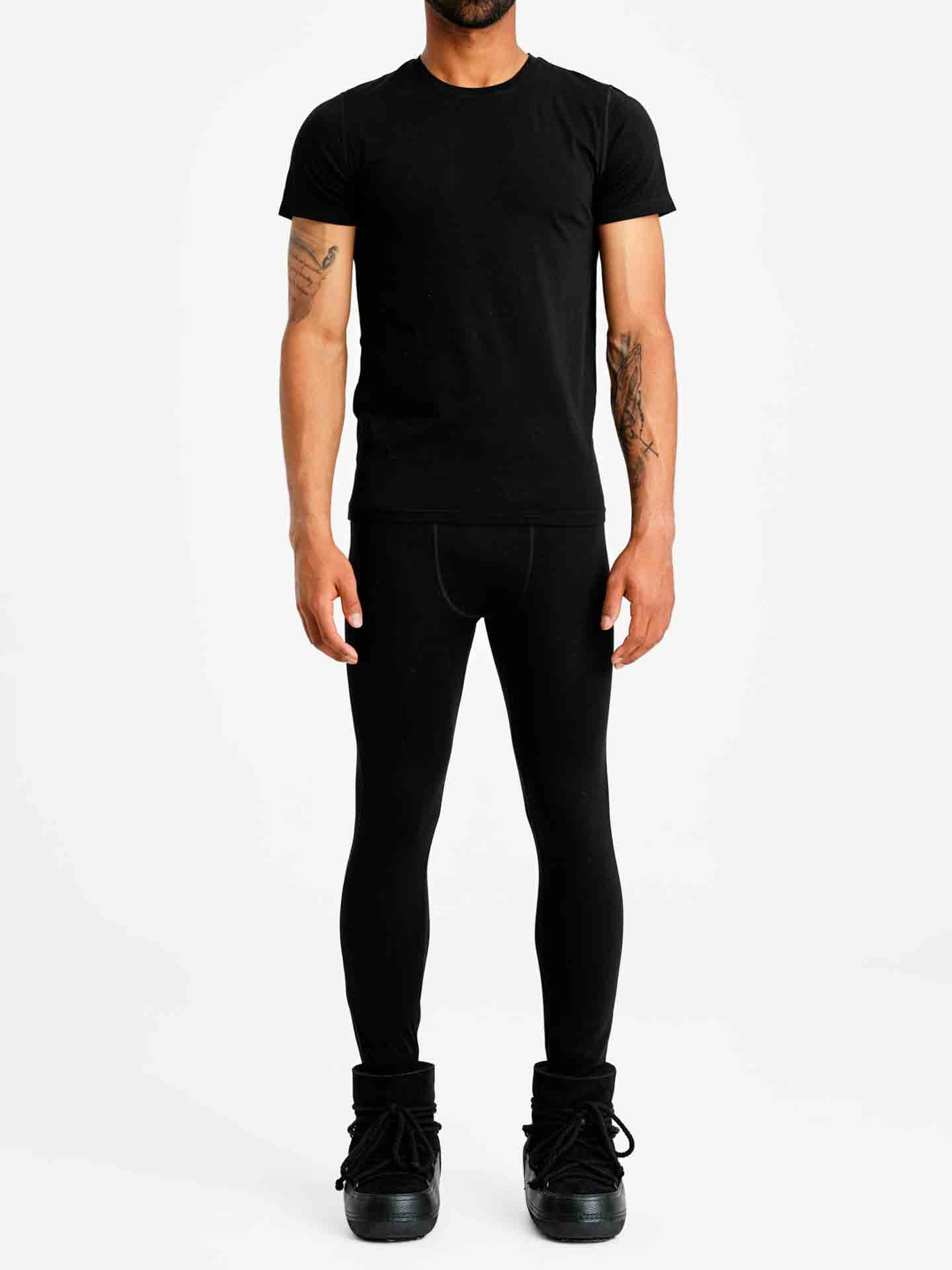A person is standing against a plain white background wearing an all-black outfit, featuring a short-sleeved Sno Merino Tee Men by We Norwegians, fitted leggings, and oversized black shoes. Their arms are relaxed by their sides.