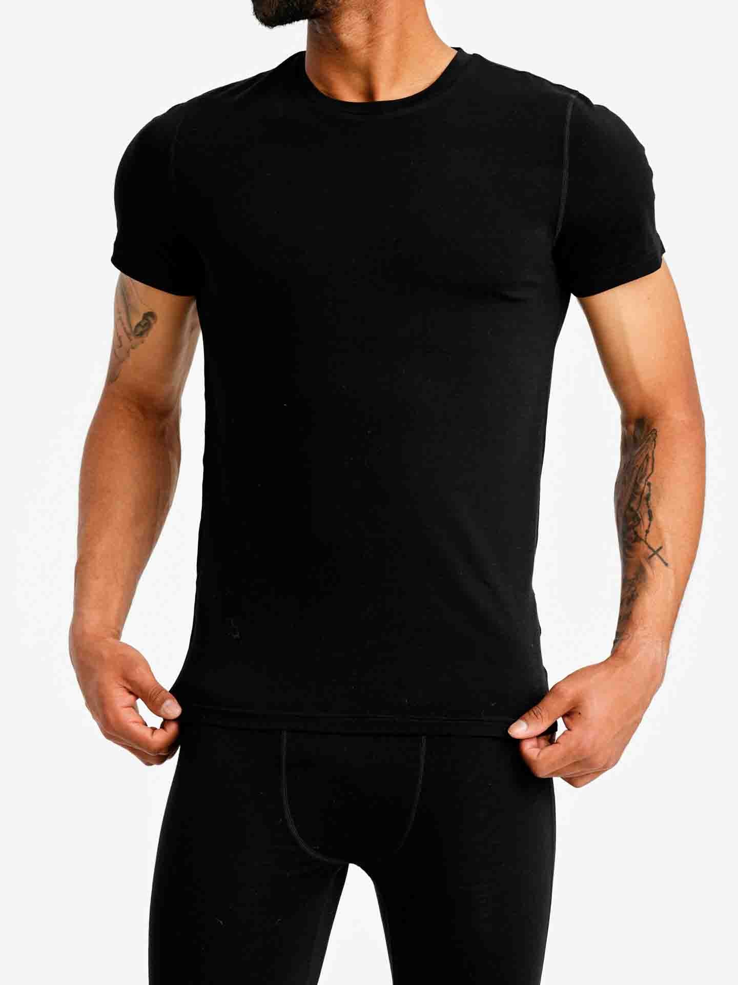 A person is wearing the We Norwegians Sno Merino Tee Men, a fitted black t-shirt made from merino wool, paired with black pants. They are holding the hem of the t-shirt with both hands. Their face is not visible, and they have tattoos on both forearms. The background is plain white.