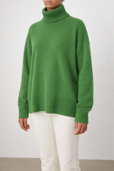A person wears the We Norwegians Blefjell Sweater Women—Pure Cashmere in green, styled with white pants, standing against a plain light background.