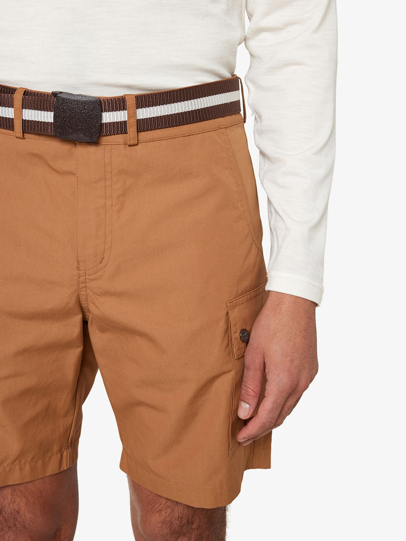 A person wearing a white long-sleeve shirt, tan shorts with a buttoned pocket, and the We Norwegians Vidda Belt featuring two white stripes around the waist stands against a plain background. Only the person's lower torso and right arm are visible.