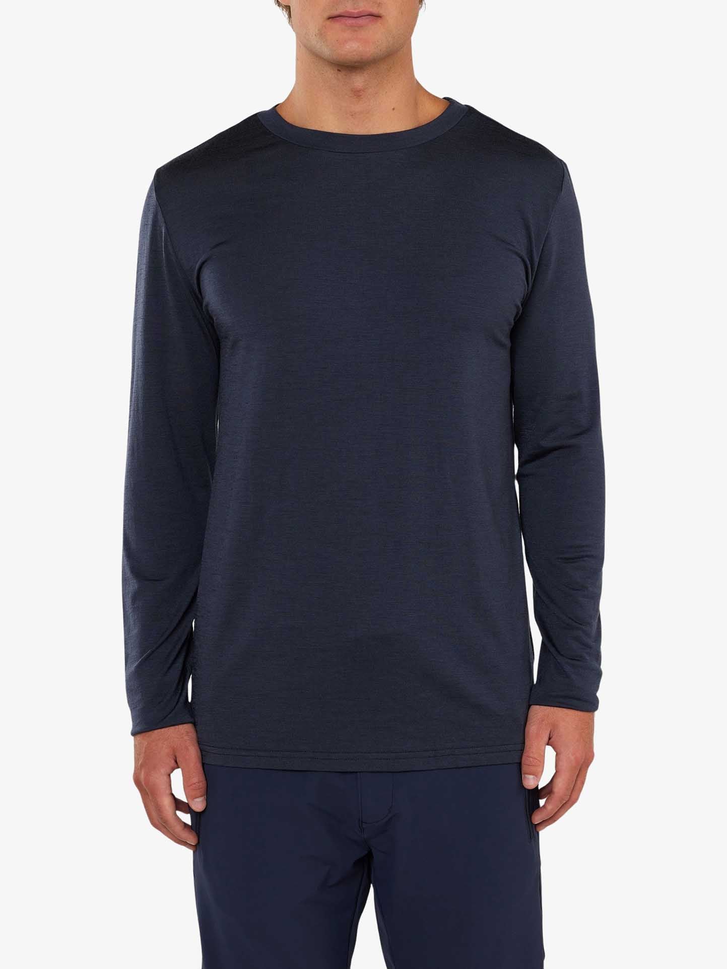 A person wearing a We Norwegians Berg LongSleeve Men, a long-sleeved shirt made from dark blue Merino wool, and matching dark blue pants stands against a plain white background. The shirt features a simple crew neckline and boasts a clean, fitted design. The person's arms are relaxed at their sides.