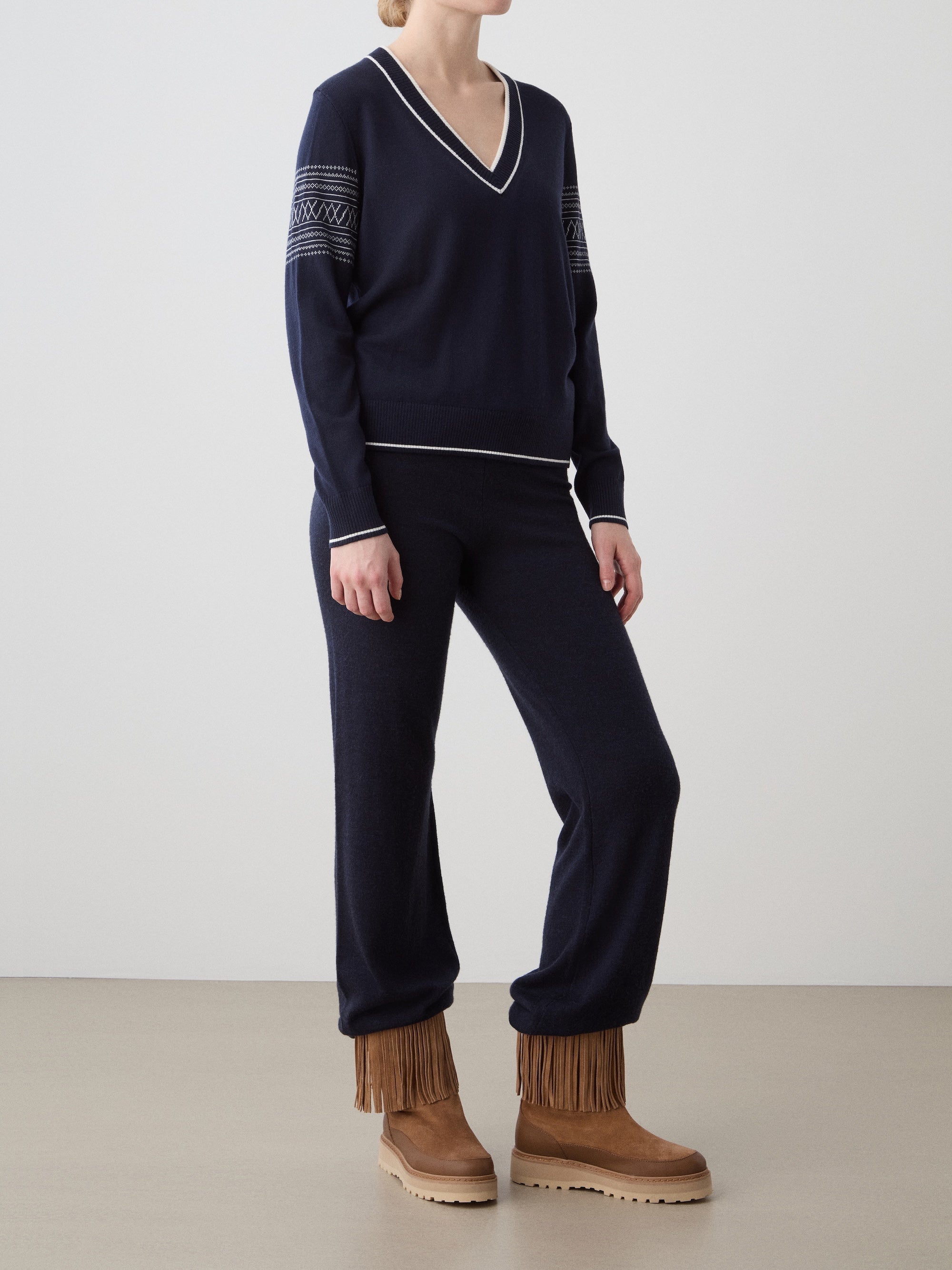 We Norwegians | FW25 | Womens Signature V-neck Sweater_Navy blue | Model wearing this fine knitted sweater with white edges on the neckline, sleeves and hemline, with the signature setesdal pattern on the sleeves - paired with the navy blue Geilo joggers and brown fringed boots | Shot against an off-white background | Front