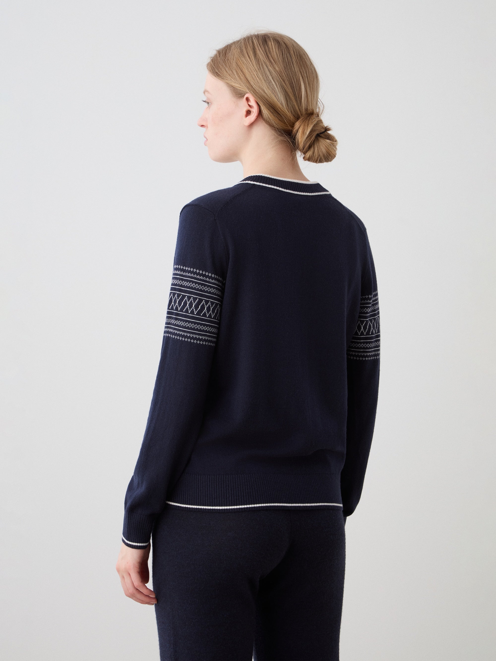 We Norwegians | FW25 | Womens Signature V-neck Sweater_Navy blue | Model wearing this fine knitted sweater with white edges on the neckline, sleeves and hemline, with the signature setesdal pattern on the sleeves - paired with the navy blue Geilo joggers | Shot against an off-white background | Back
