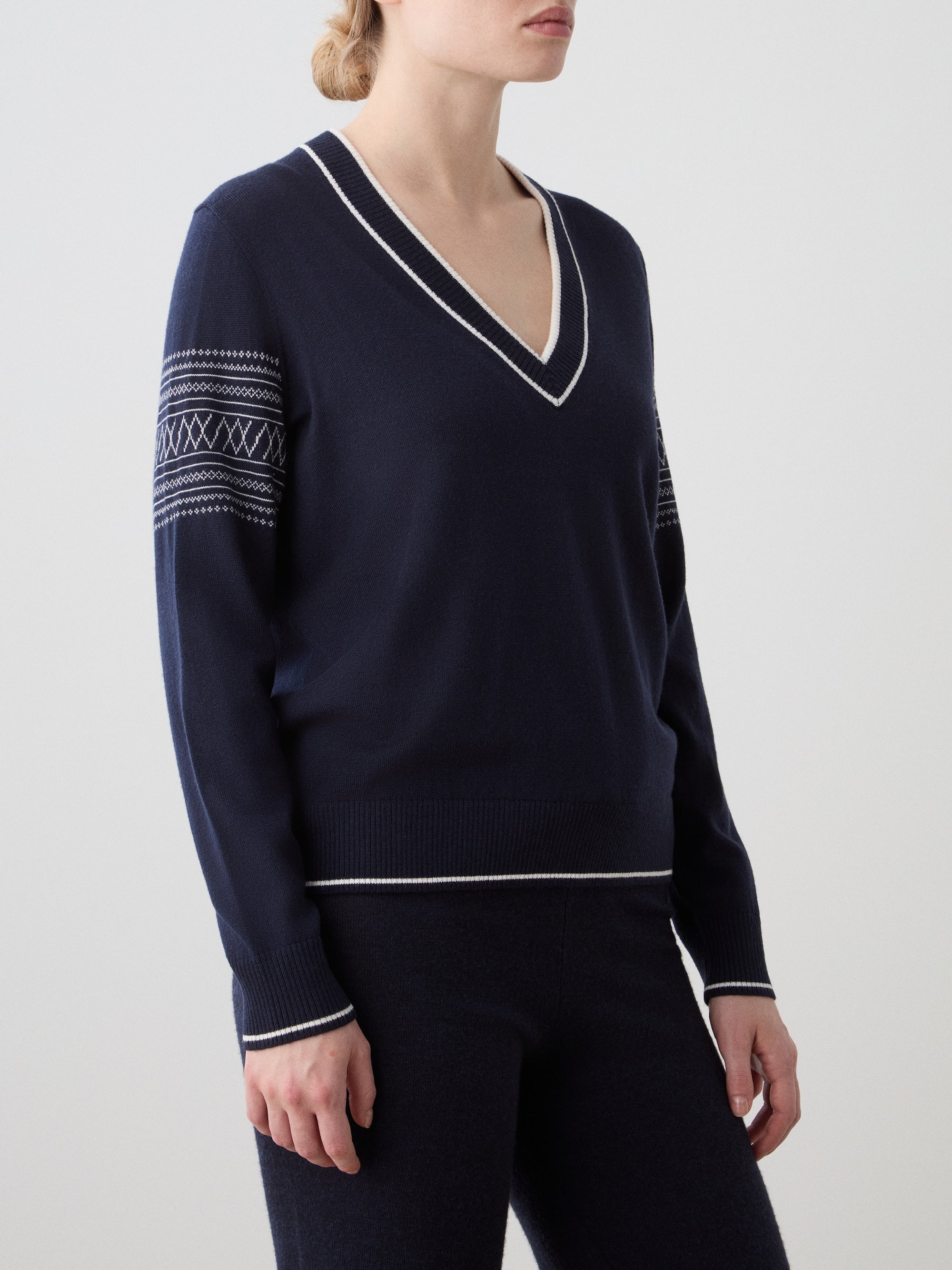 We Norwegians | FW25 | Womens Signature V-neck Sweater_Navy blue | Model wearing this fine knitted sweater with white edges on the neckline, sleeves and hemline, with the signature setesdal pattern on the sleeves - paired with the navy blue Geilo joggers | Shot against an off-white background | Close-up