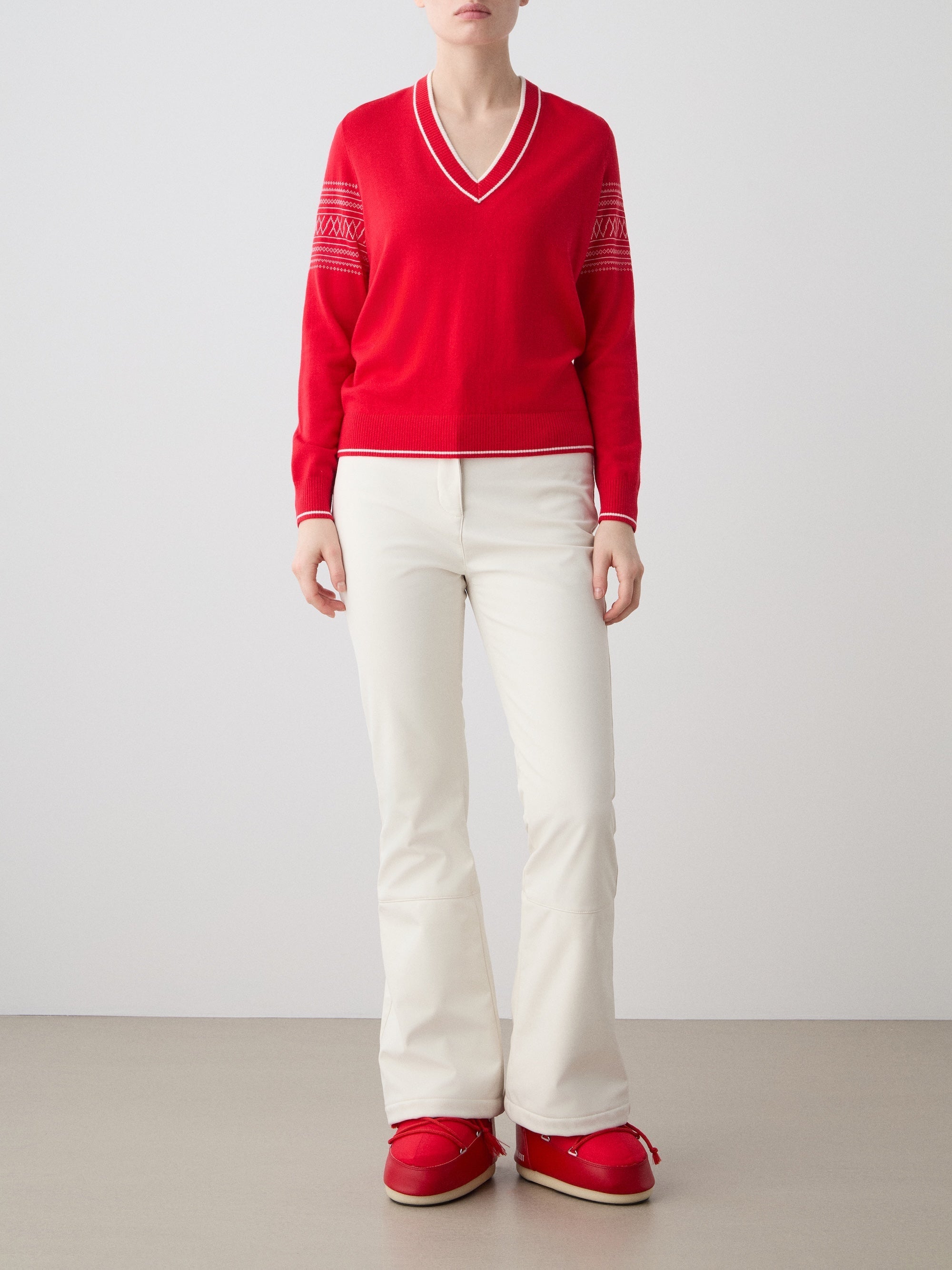 We Norwegians | FW25 | Womens Signature V-neck Sweater_Red | Model wearing this fine knitted sweater with white edges on the neckline, sleeves and hemline, with the signature setesdal pattern on the sleeves - paired with the off-white Tyin bootleg ski pants and red snow boots | Shot against an off-white background | Front