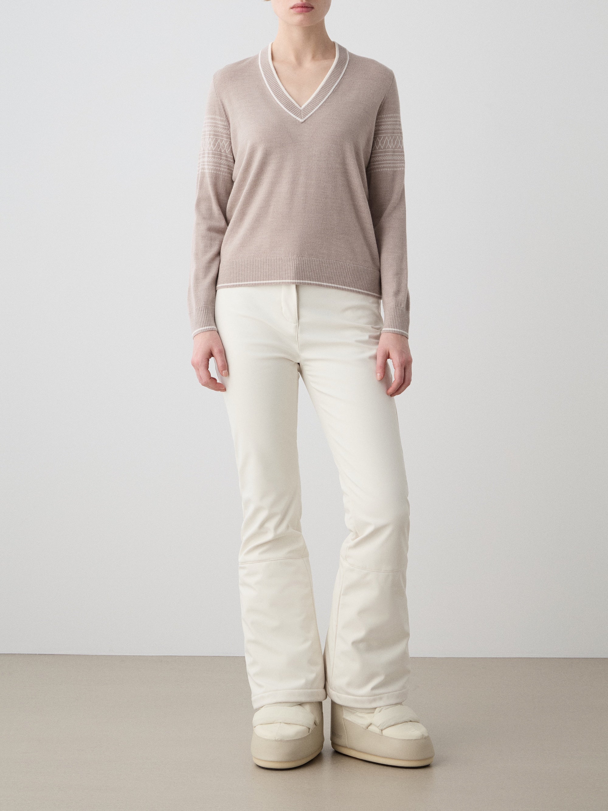 We Norwegians | FW25 | Womens Signature V-neck Sweater_Sand | Model wearing this fine knitted sweater with white edges on the neckline, sleeves and hemline, with the signature setesdal pattern on the sleeves - paired with the off-white Tyin bootleg ski pants and white snow boots | Shot against an off-white background | Front