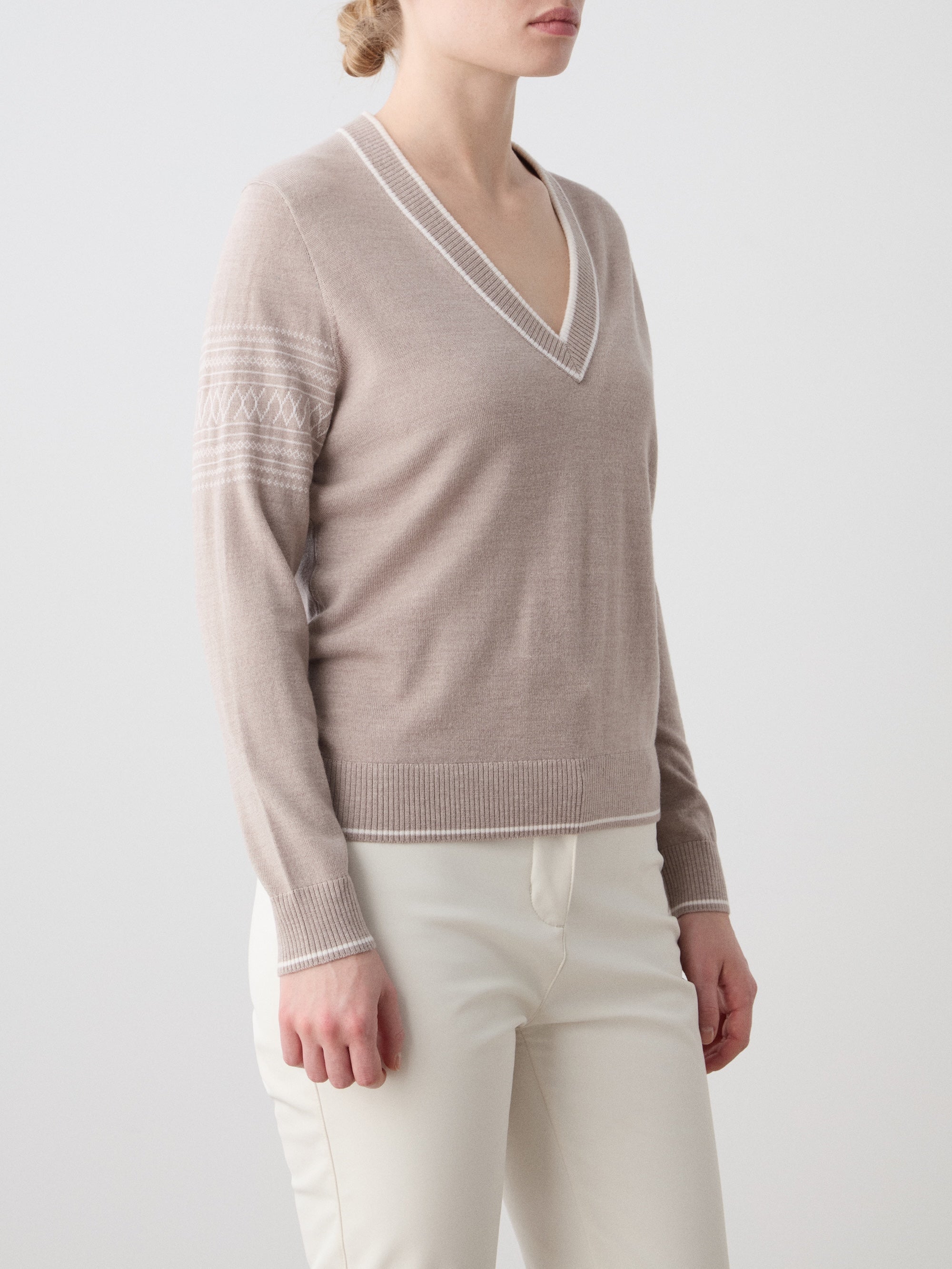We Norwegians | FW25 | Womens Signature V-neck Sweater_Sand | Model wearing this fine knitted sweater with white edges on the neckline, sleeves and hemline, with the signature setesdal pattern on the sleeves - paired with the off-white Tyin bootleg ski pants | Shot against an off-white background | Close-up