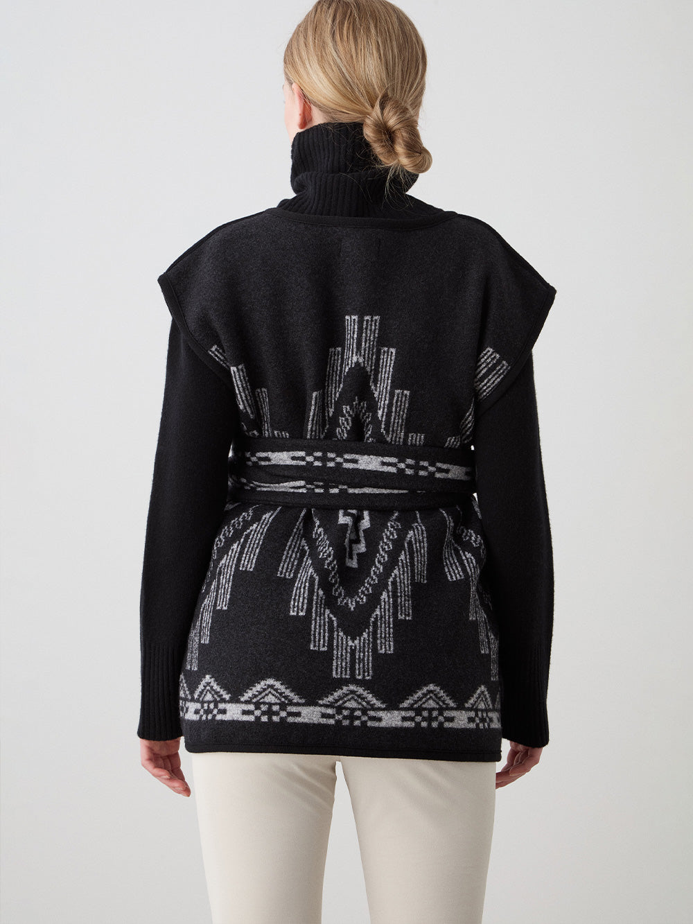 We Norwegians_FW25_Womens | Model is wearing the Kautokeino vest | Black in color with a jacquard pattern inspired by Native and Indigenous American Art | Vest has a matching belt to cinch the waist | Back view on a white background