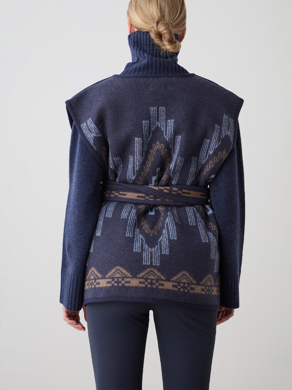 We Norwegians_FW25_Womens | Model is wearing the Kautokeino vest | Navy blue in color with a jacquard pattern inspired by Native and Indigenous American Art | Vest has a matching belt to cinch the waist | Back view on a white background