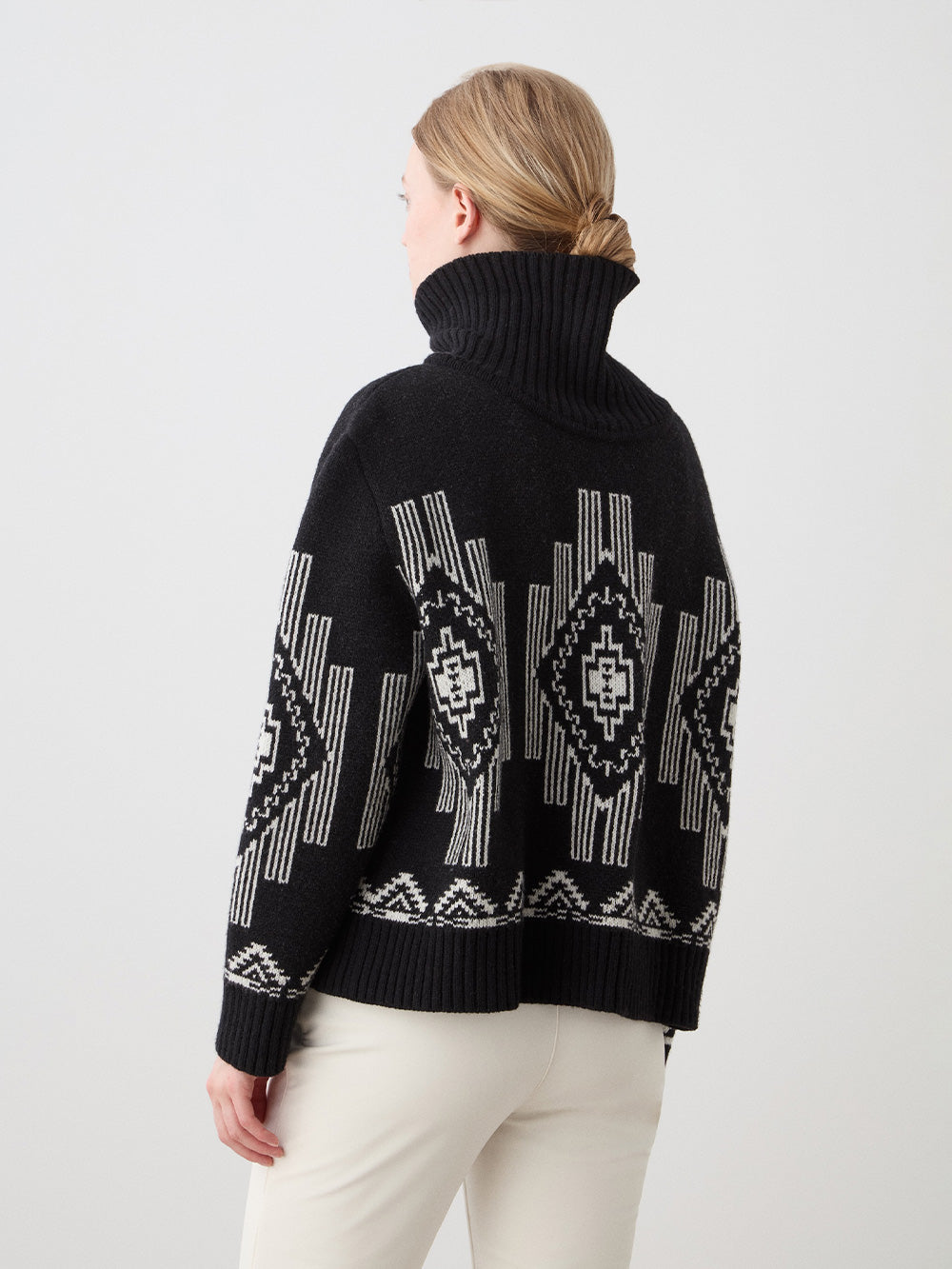 A person with light hair in a low bun is seen from the back, wearing the We Norwegians Kautokeino Sweater women—a black wool turtleneck with white geometric patterns—paired with cream pants against a plain light background.