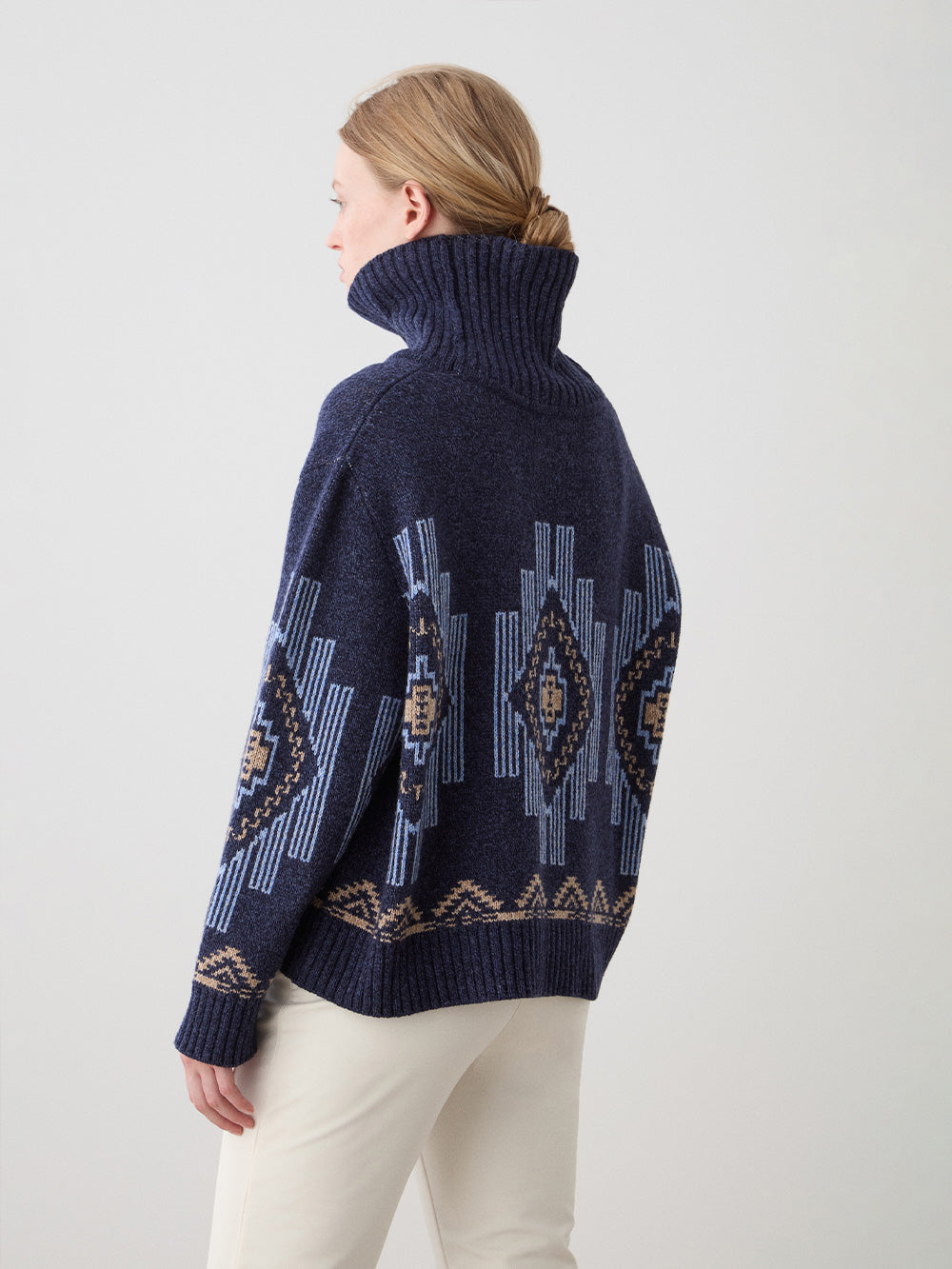A light-haired woman models the We Norwegians Kautokeino Sweater in indigo with geometric patterns and cream pants, standing against a plain light background, shown from the back and side.