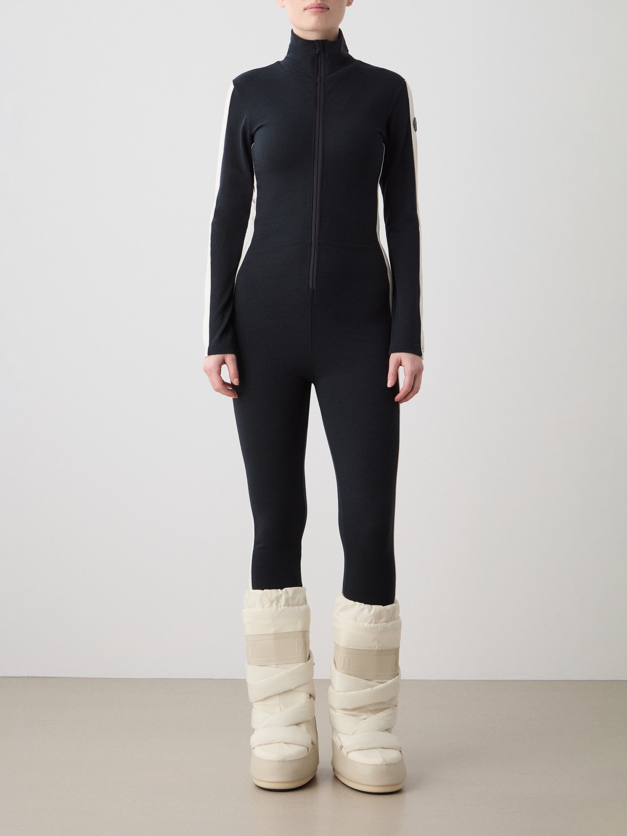 We Norwegians | FW25 | Womens Voss Jumpsuit_Black | The model is wearing this piqué knit jumpsuit with a front zipper opening, a stand collar, white panels on the sleeves with subtle branding - paired with white snow boots | Shot against an off-white background | Front