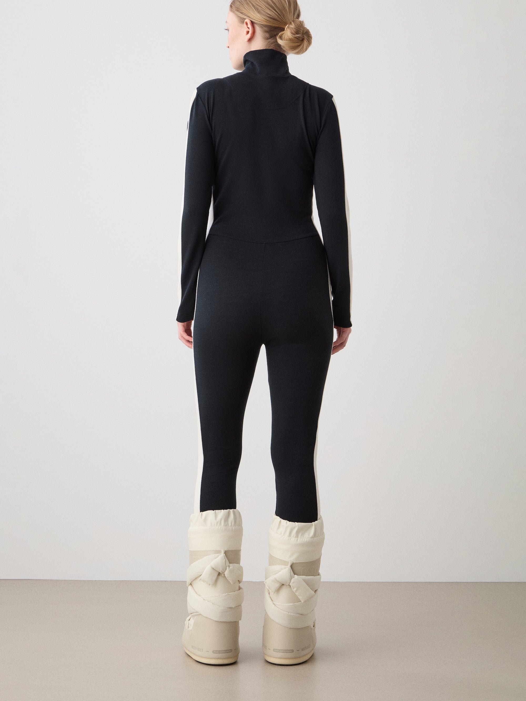 We Norwegians | FW25 | Womens Voss Jumpsuit_Black | The model is wearing this piqué knit jumpsuit with a front zipper opening, a stand collar, white panels on the sleeves with subtle branding - paired with white snow boots | Shot against an off-white background | Back