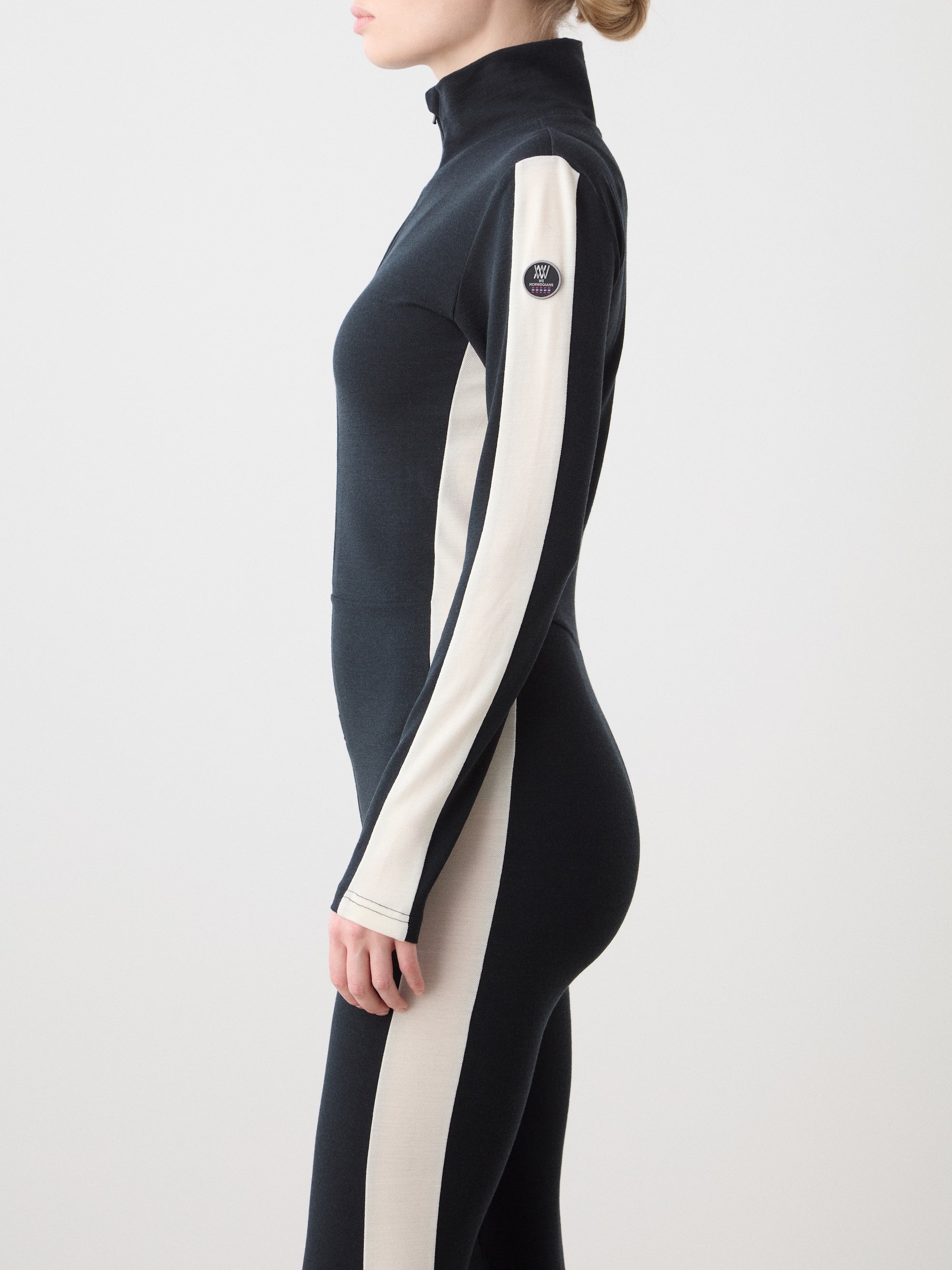We Norwegians | FW25 | Womens Voss Jumpsuit_Black | The model is wearing this piqué knit jumpsuit with a front zipper opening, a stand collar, white panels on the sleeves with subtle branding | Shot against an off-white background | Side/ Close-up