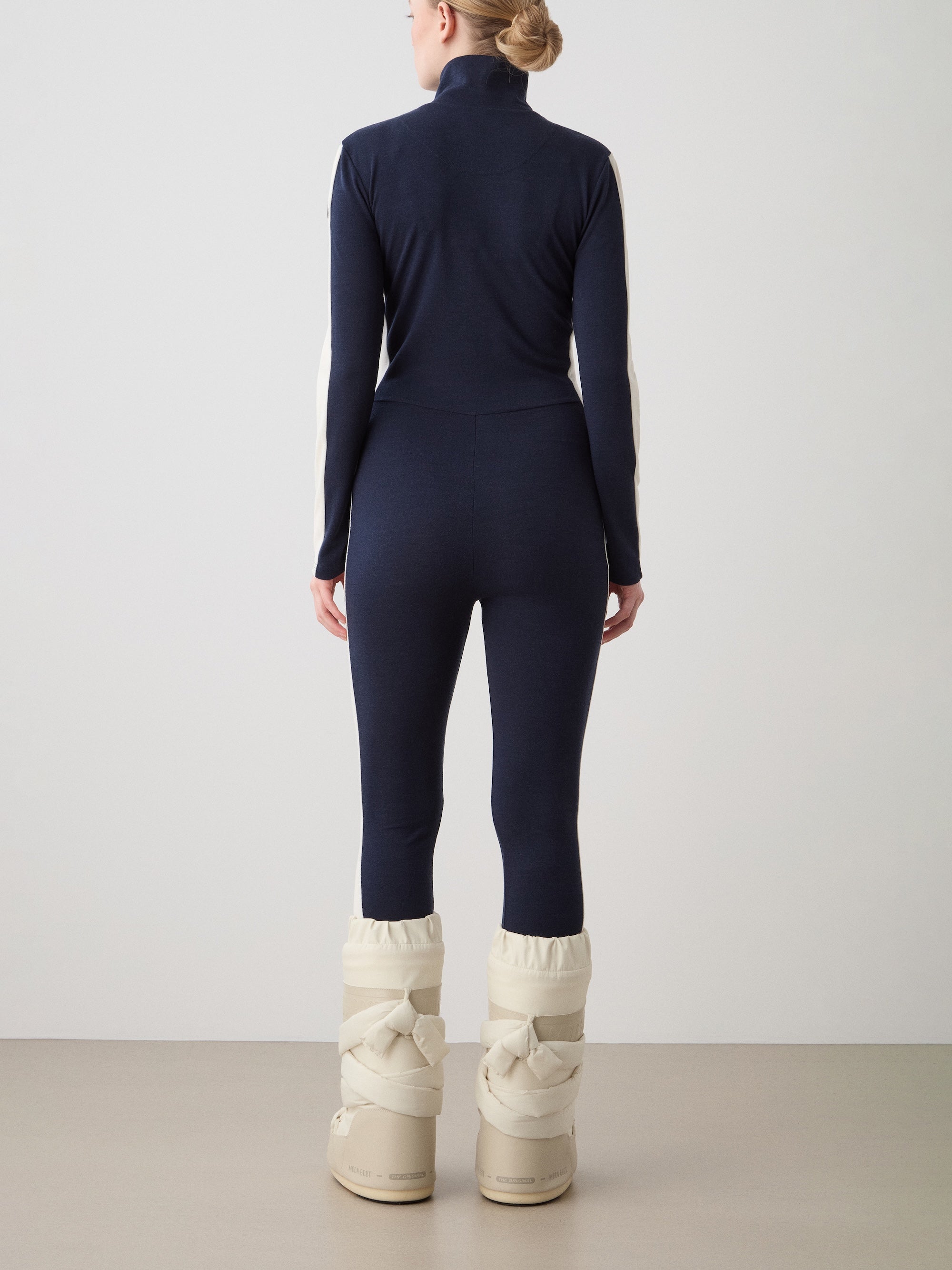 We Norwegians | FW25 | Womens Voss Jumpsuit_Navy blue | The model is wearing this piqué knit jumpsuit with a front zipper opening, a stand collar, white panels on the sleeves with subtle branding - paired with white snow boots | Shot against an off-white background | Back