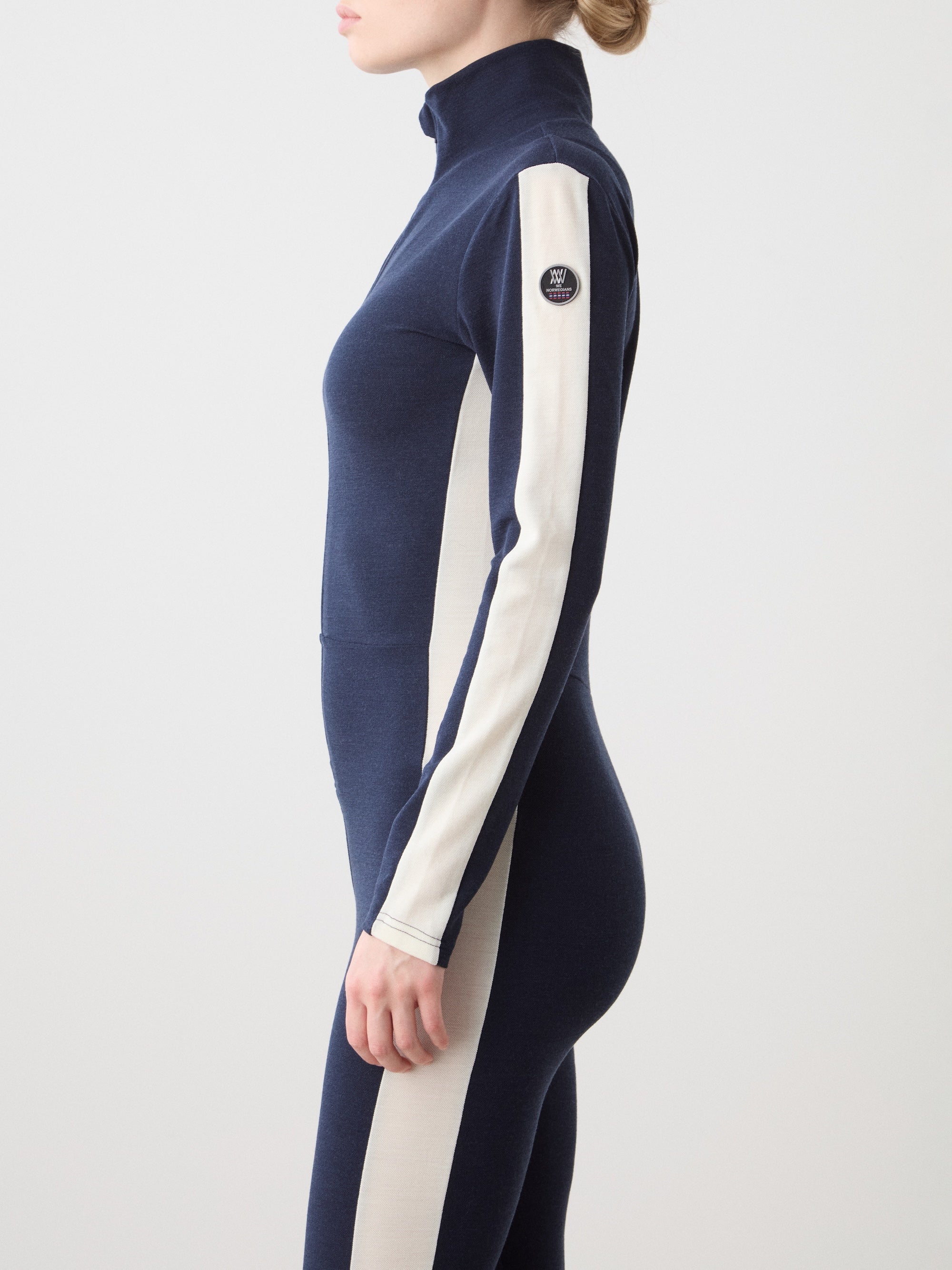 We Norwegians | FW25 | Womens Voss Jumpsuit_Navy blue | The model is wearing this piqué knit jumpsuit with a front zipper opening, a stand collar, white panels on the sleeves with subtle branding | Shot against an off-white background | Side/ Close-up