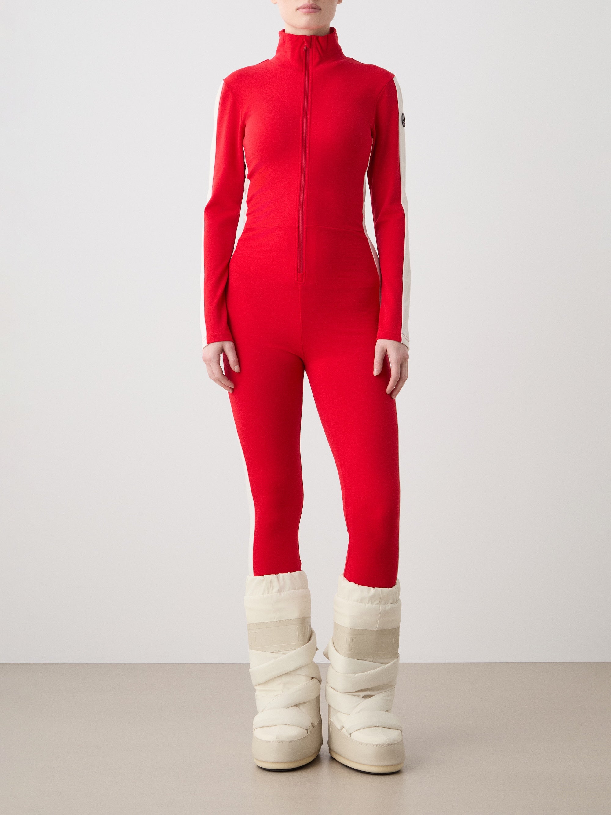 We Norwegians | FW25 | Womens Voss Jumpsuit_Red | The model is wearing this piqué knit jumpsuit with a front zipper opening, a stand collar, white panels on the sleeves with subtle branding - paired with white snow boots | Shot against an off-white background | Front