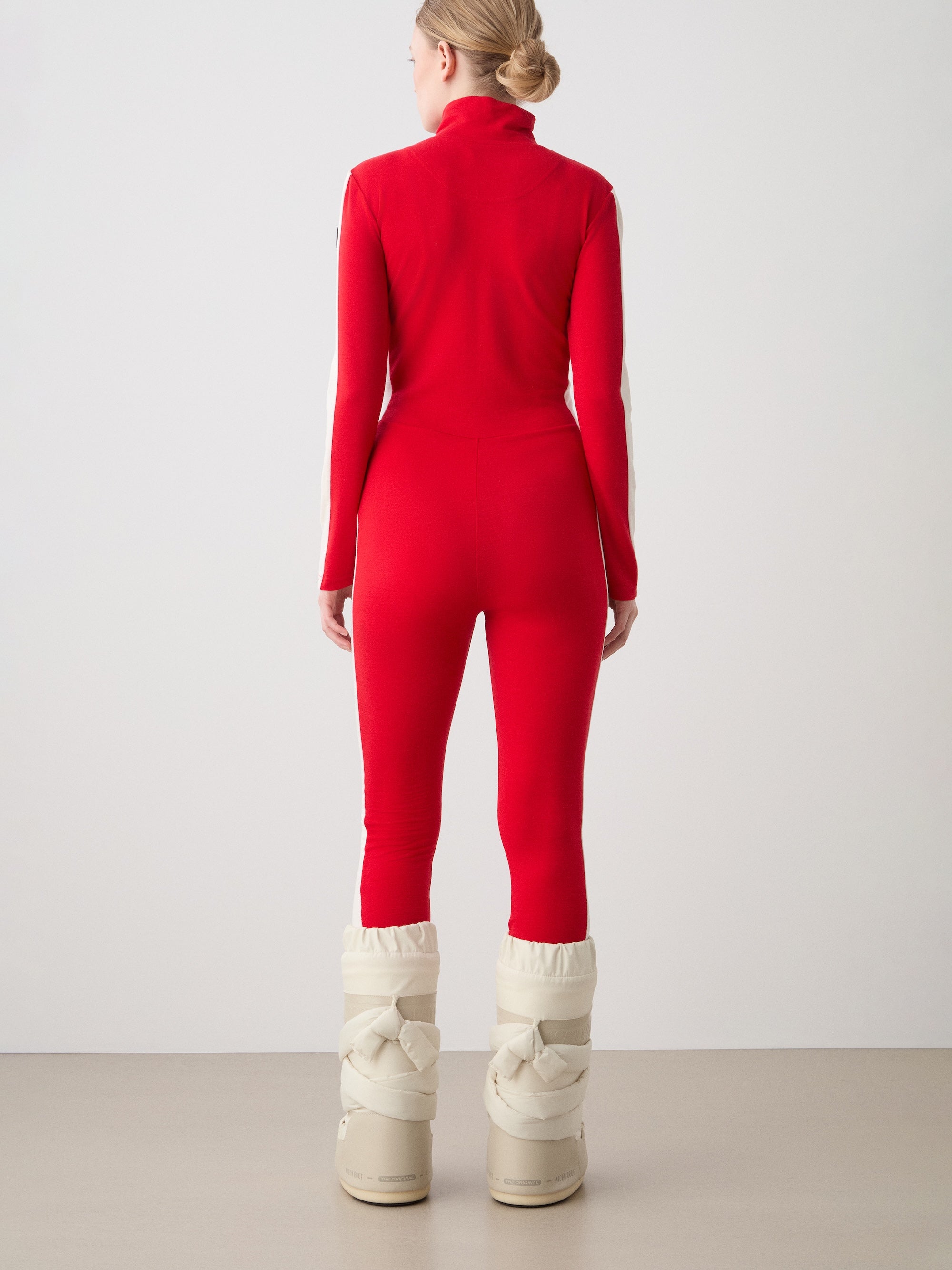 We Norwegians | FW25 | Womens Voss Jumpsuit_Red | The model is wearing this piqué knit jumpsuit with a front zipper opening, a stand collar, white panels on the sleeves with subtle branding - paired with white snow boots | Shot against an off-white background | Back