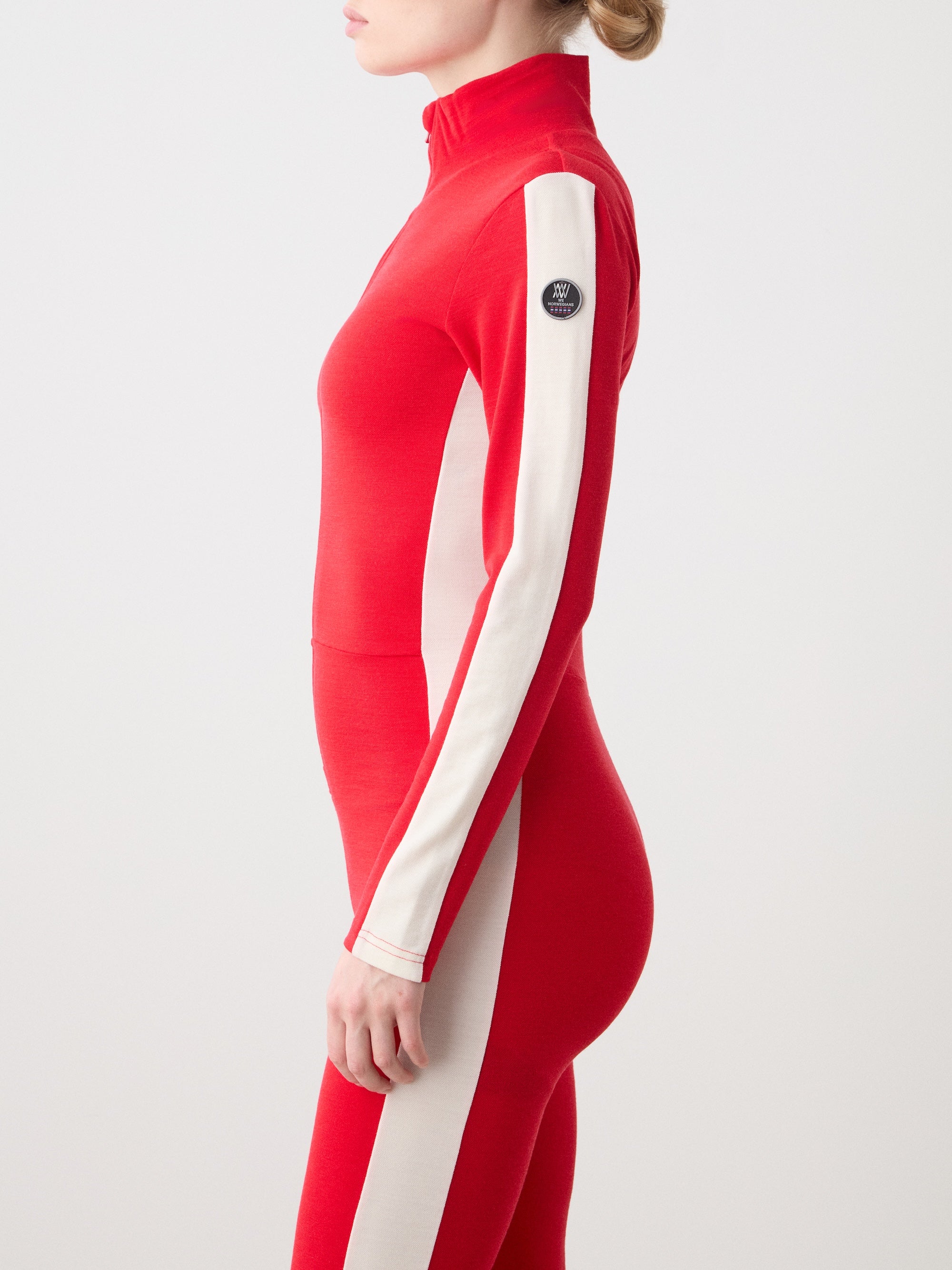 We Norwegians | FW25 | Womens Voss Jumpsuit_Red | The model is wearing this piqué knit jumpsuit with a front zipper opening, a stand collar, white panels on the sleeves with subtle branding | Shot against an off-white background | Side/ Close-up