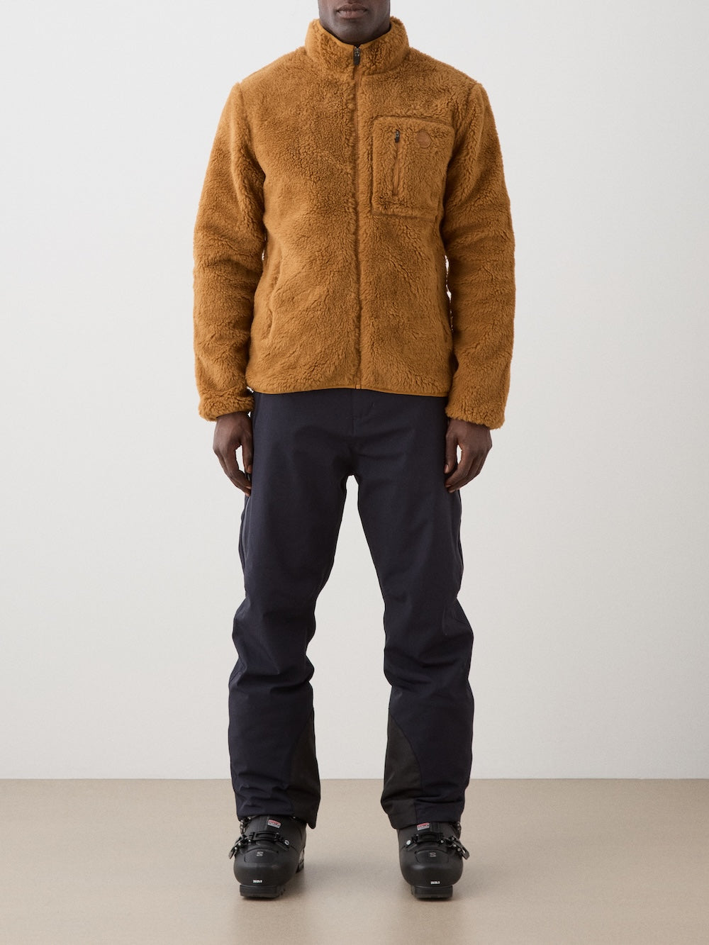 We Norwegians | FW25 | Mens Kebnekaise Jacket_Cinnamon | Model wearing this fleece jacket with front zipper opening, zipped chest pocket and side pockets - paired with black ski pants | Shot against an off-white background | Front