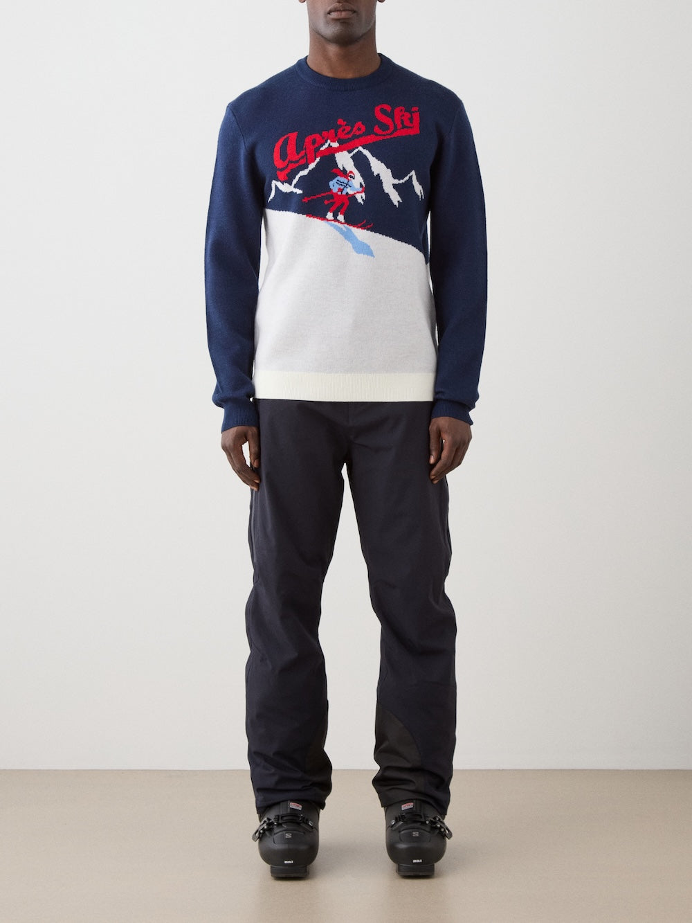 We Norwegians | FW25 | Mens Afterski Crewneck_Navy blue | Model wearing a knitted sweater with an après ski design at the front with black ski pants | Shot against an off-white background | Front view