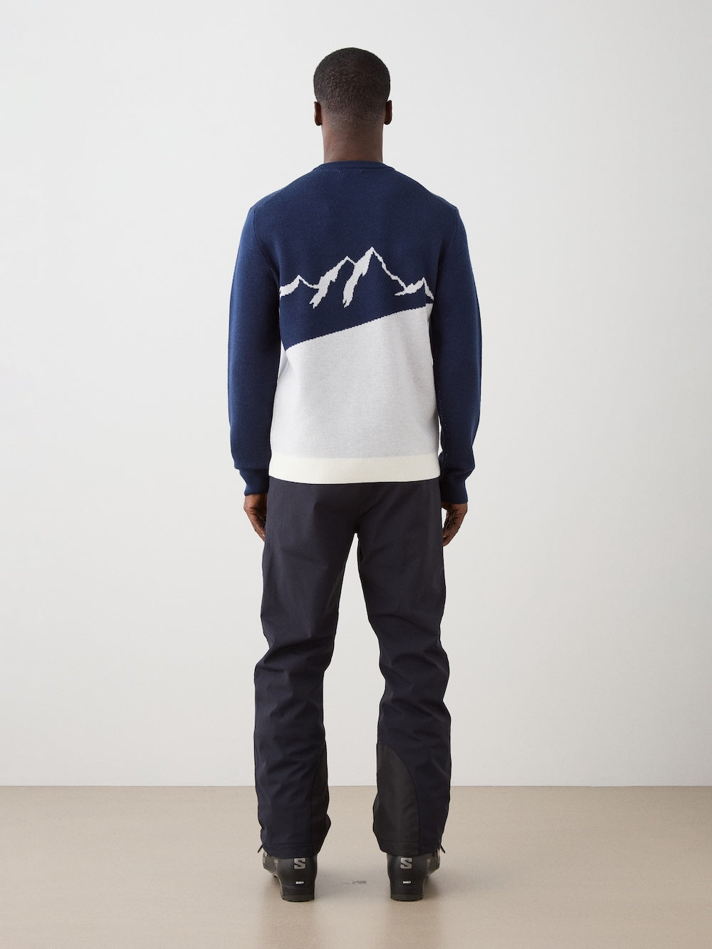 We Norwegians | FW25 | Mens Afterski Crewneck_Navy blue | Model wearing a knitted sweater with an après ski design at the front with black ski pants | Shot against an off-white background | Back view