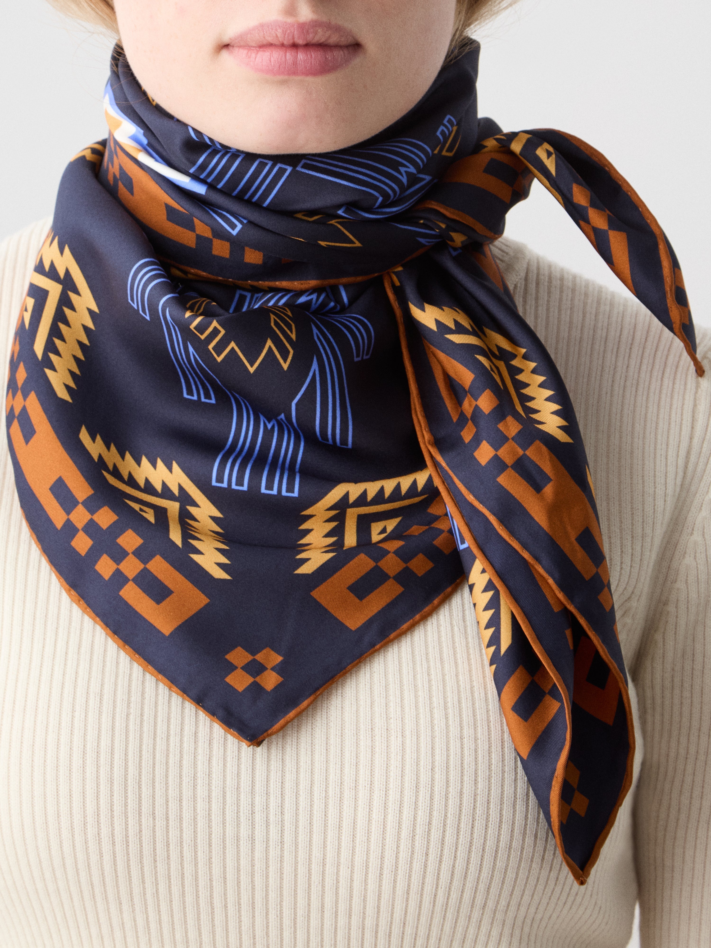 We Norwegians | FW25 | Pioneer Silk Scarf_Navy blue | Female model wearing the scarf with printed motifs over a light top | Shot against an off-white background