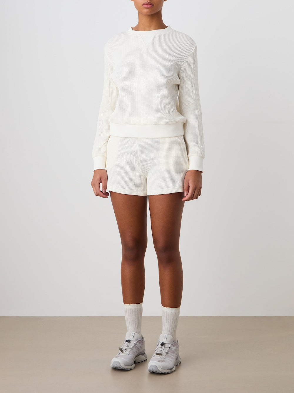 Person wearing an off white waffle knit crewneck with matching shorts, white socks and sneakers against a plain background