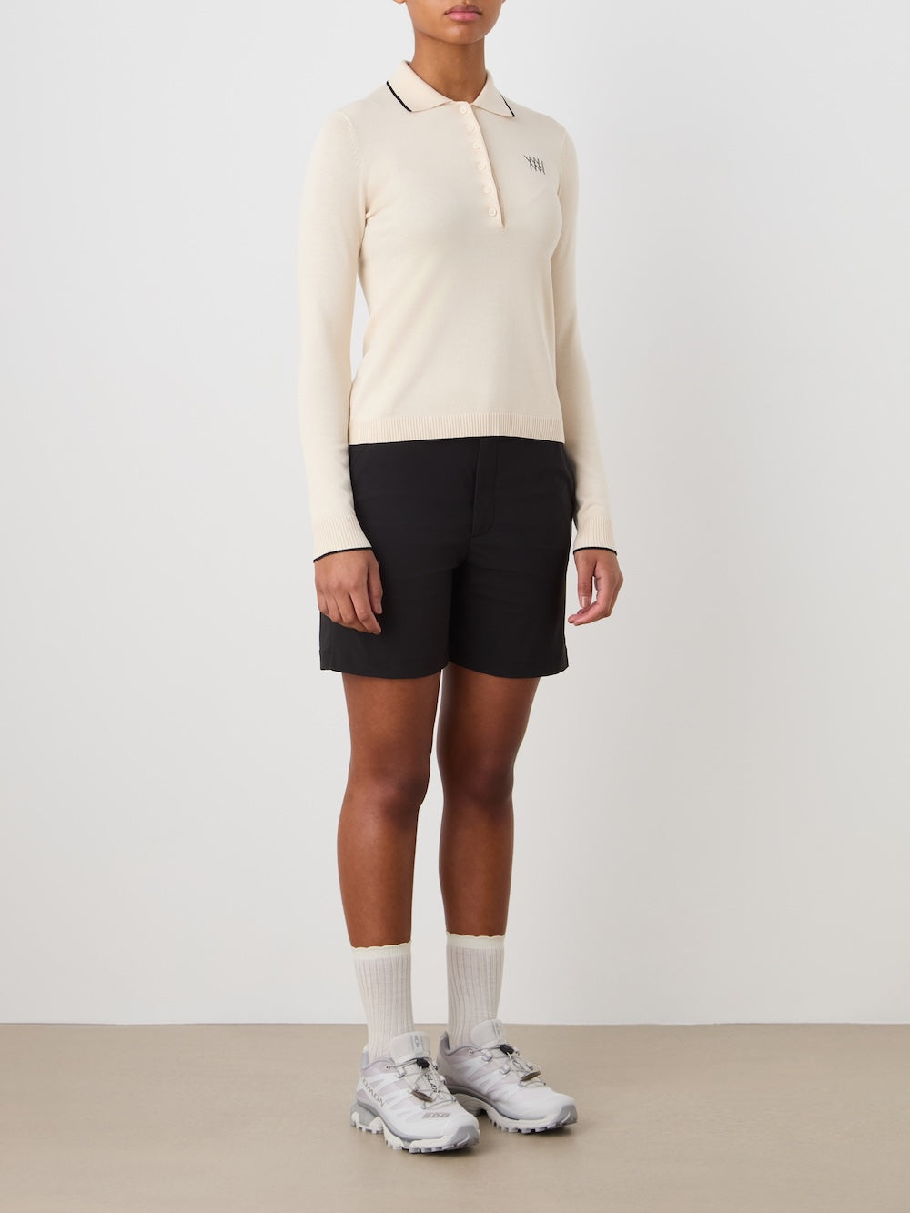 Person wearing a beige long-sleeve polo, black shorts, and white sneakers on a plain background