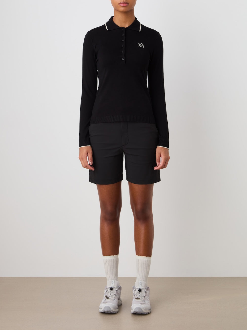 Person wearing a black long-sleeve polo and shorts with white socks and shoes on a plain background