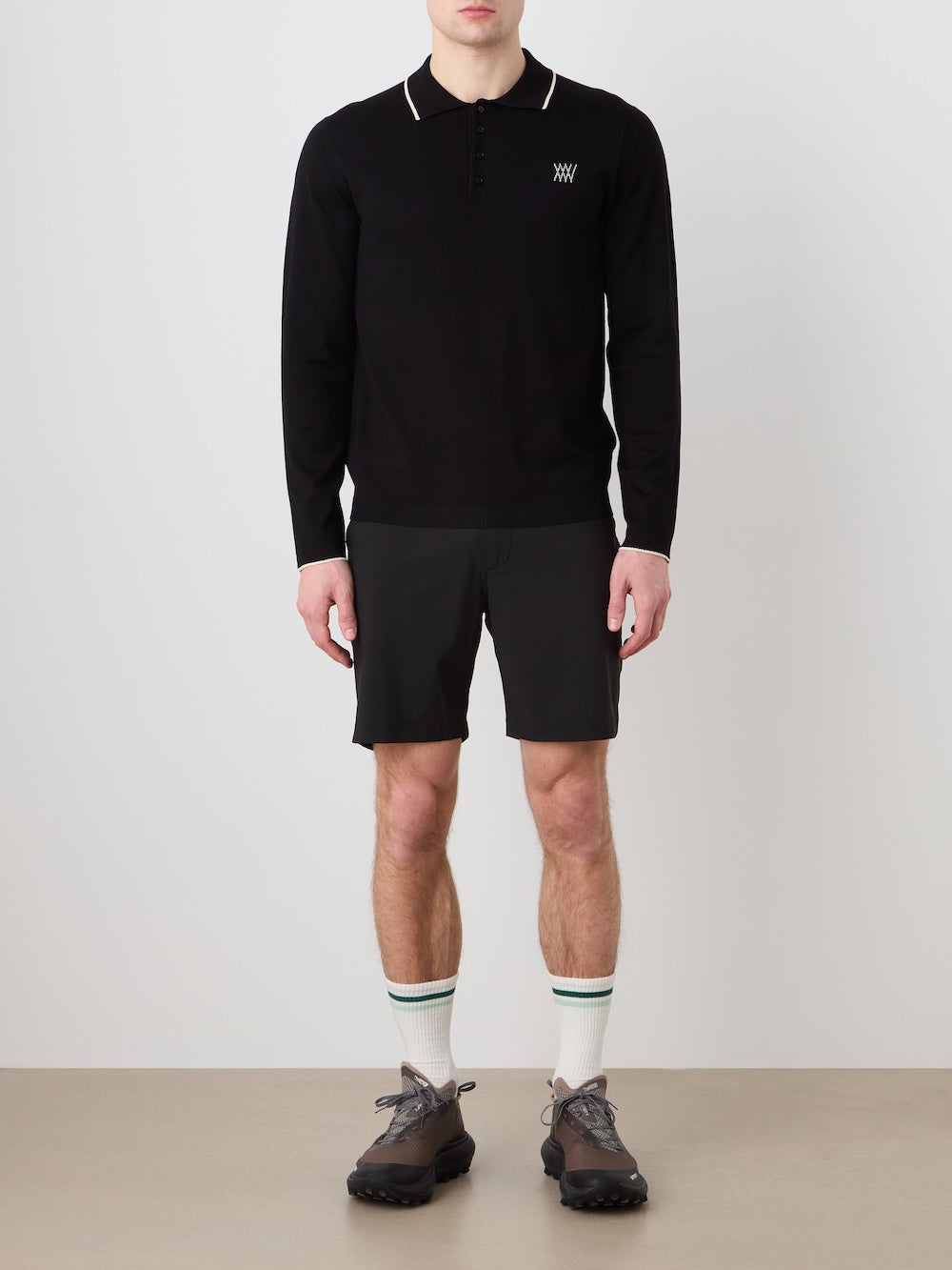 Person wearing a black long sleeved polo, shorts, and shoes with white socks against a plain background