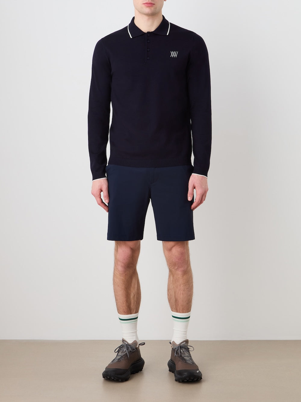 Person wearing a navy blue long sleeved polo, shorts, and shoes against a light background