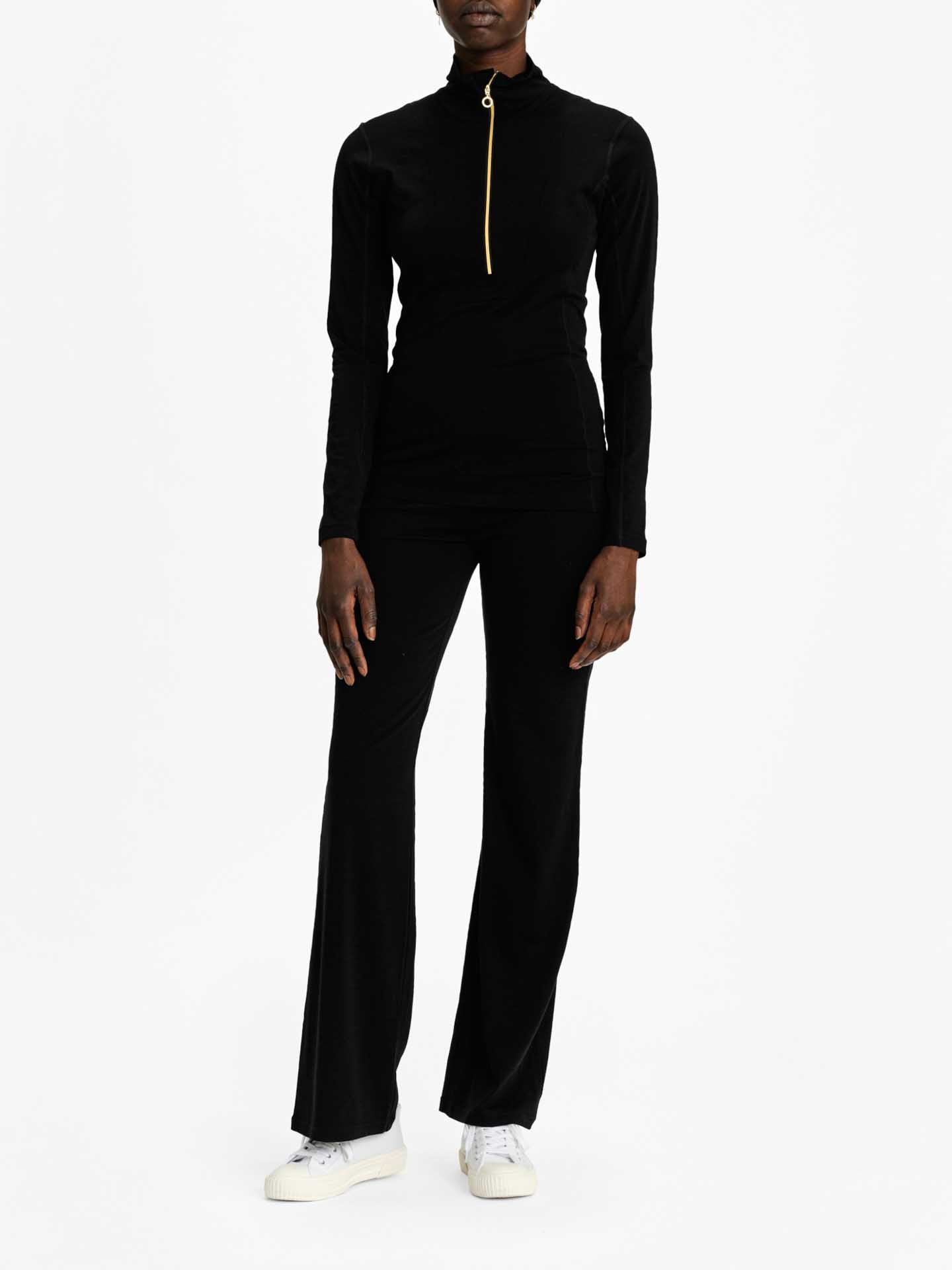 A person stands against a plain white background, wearing the Sno Merino Zip Up Women by We Norwegians, a fitted black long-sleeve top featuring a gold zipper running from the collar down to the chest, paired with matching black flared pants. They are also wearing white sneakers. The person's hands rest gently at their sides.
