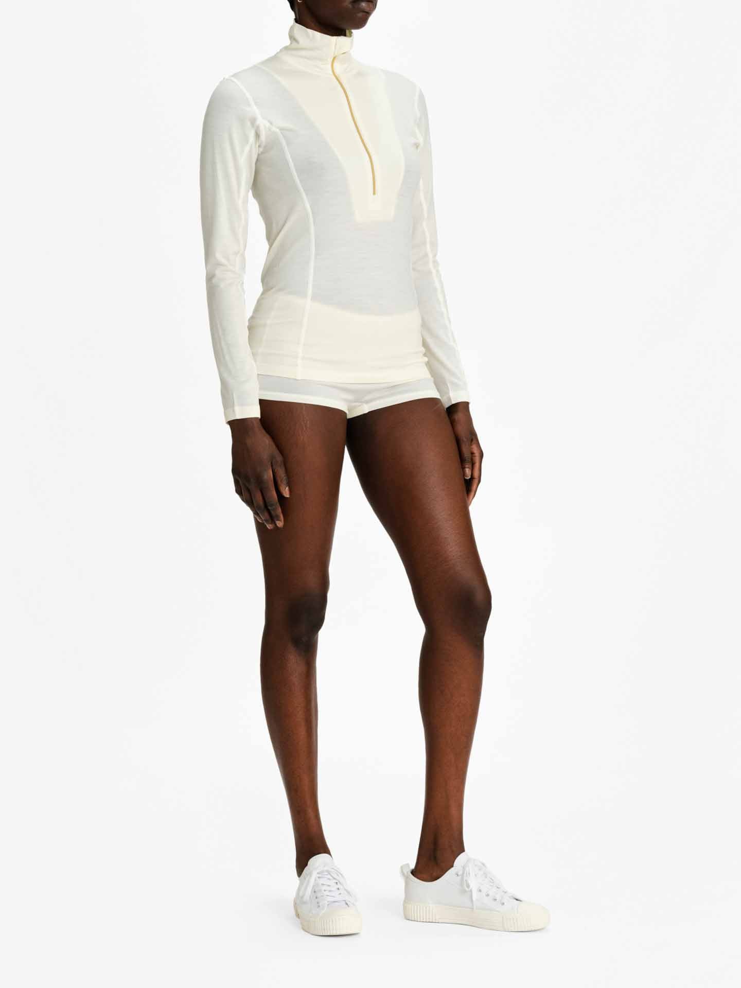 A person is wearing the Sno Merino Zip Up Women, a fitted, long-sleeved white athletic top with a high collar and quarter zipper from We Norwegians' luxury Scandinavian base-layers collection. Paired with form-fitting white shorts and white sneakers, they stand against a plain white background.