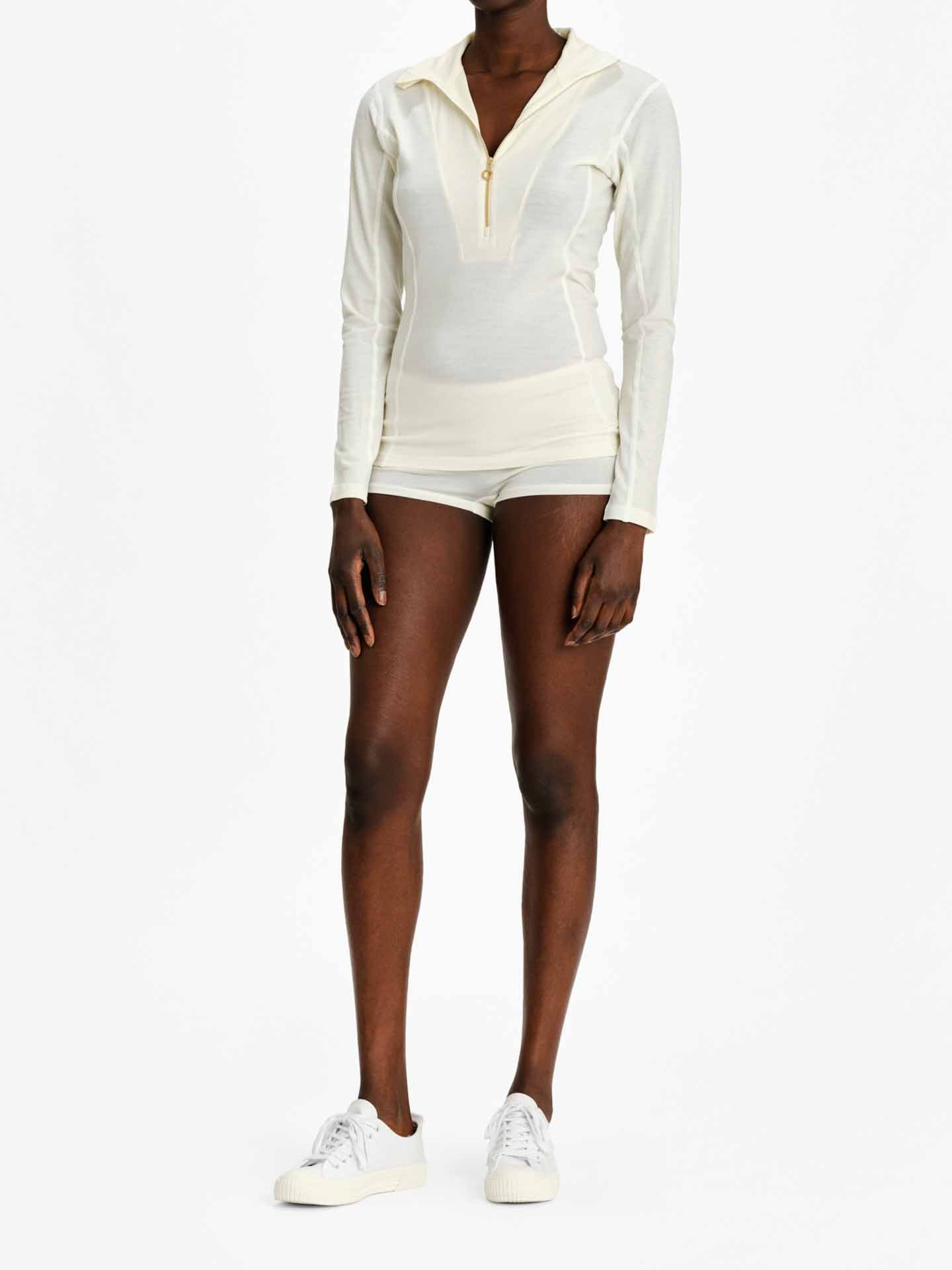 A person is wearing the Sno Merino Zip Up Women by We Norwegians, a light-colored, long-sleeve athletic top with a fitted shape and quarter-zip neckline, crafted from super fine merino wool. Matching short shorts complete the look as they stand against a plain white background, complemented by white sneakers.