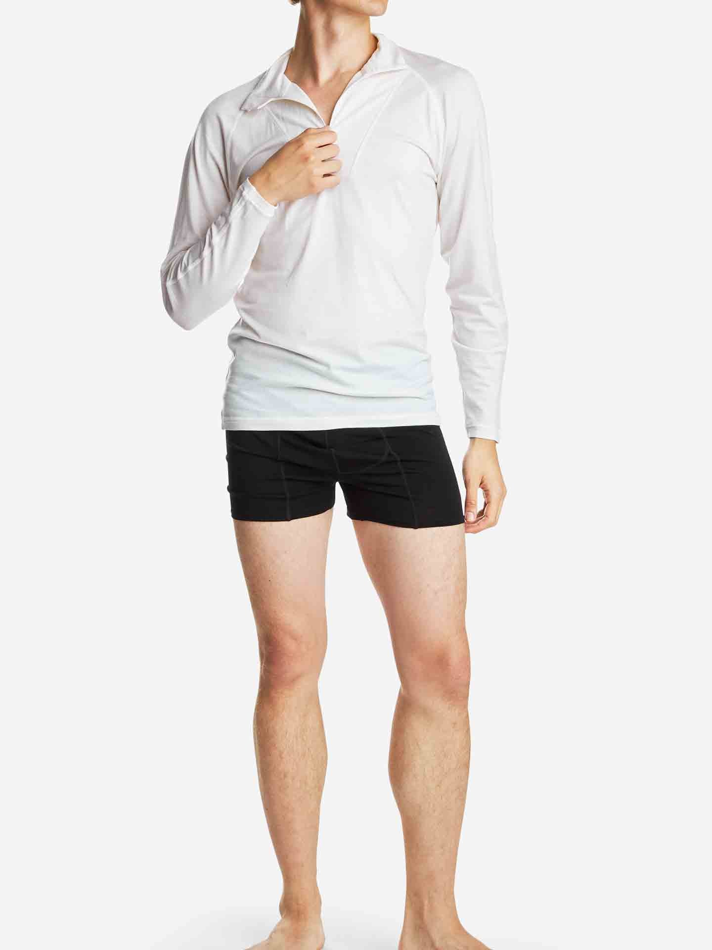 A person standing against a white background is wearing the Sno Merino Zip Up Men by We Norwegians. They are barefoot and holding the collar of their long-sleeved, white merino wool shirt with one hand. Paired with black shorts, this simple, casual outfit highlights the Scandinavian luxury base-layer known for its breathability.