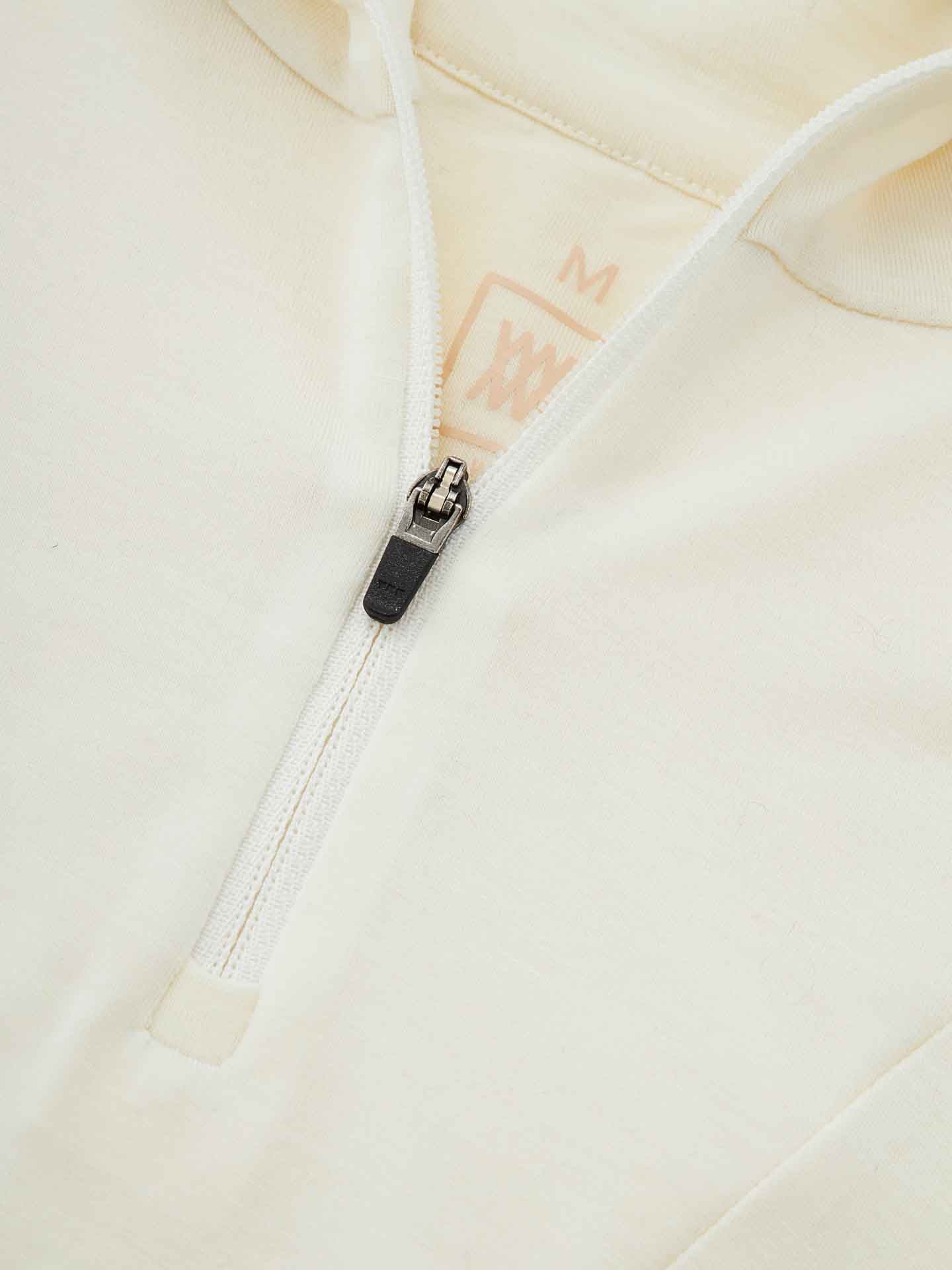 Close-up image of the We Norwegians Sno Merino Zip Up Men in a beige or off-white color, featuring a front zipper. The black and silver zipper pull is partially unzipped. A size "M" label is visible at the collar. Made from breathable merino wool, the material appears soft and comfortable, epitomizing Scandinavian luxury base-layers.