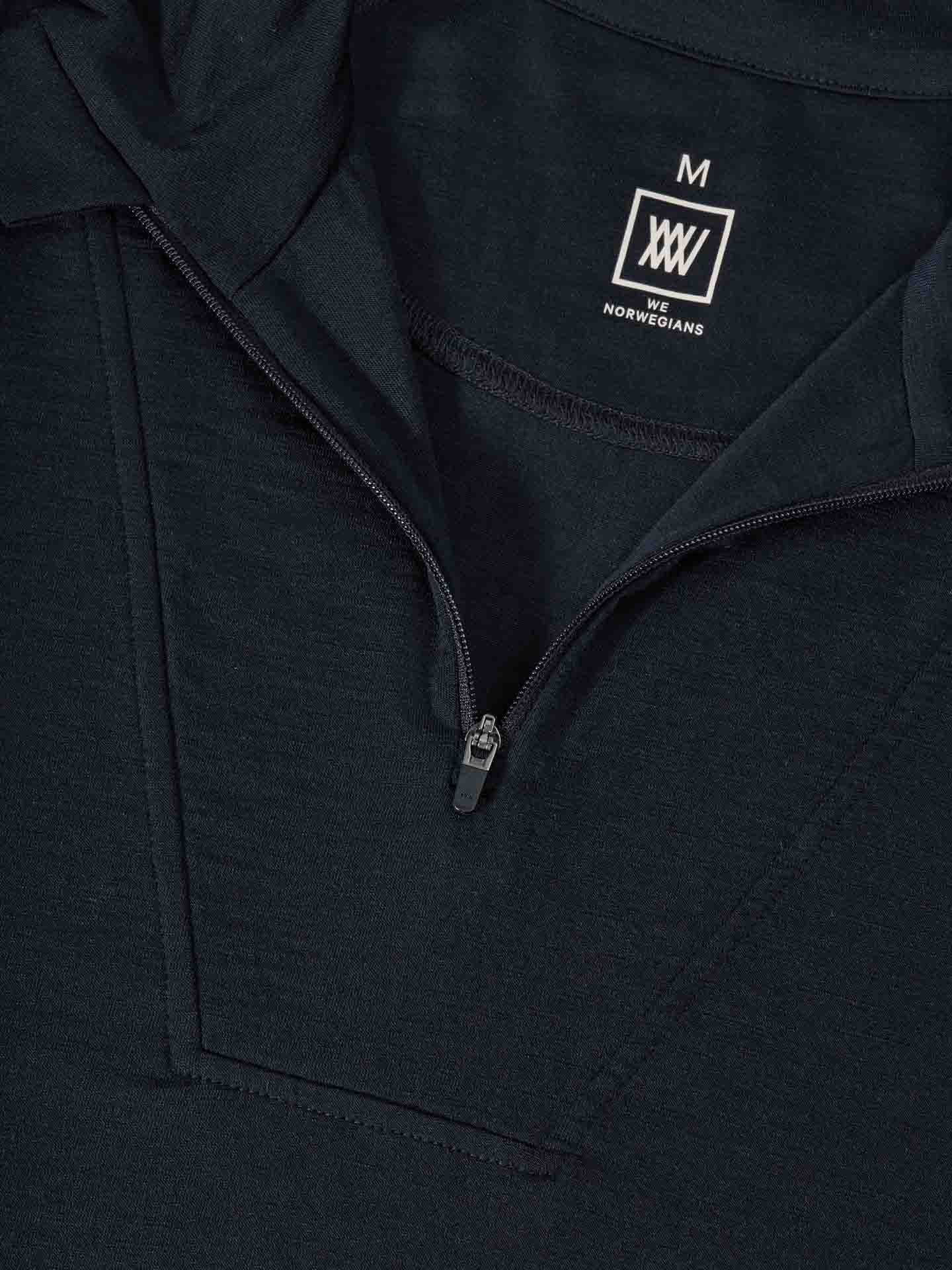 Close-up of the Sno Merino Zip Up Men in a dark color, featuring a half-zip front. The zipper is partially open, revealing a glimpse of the breathable merino wool fabric underneath. A label inside the garment reads "M" and includes the logo for We Norwegians, known for their Scandinavian luxury base-layers.