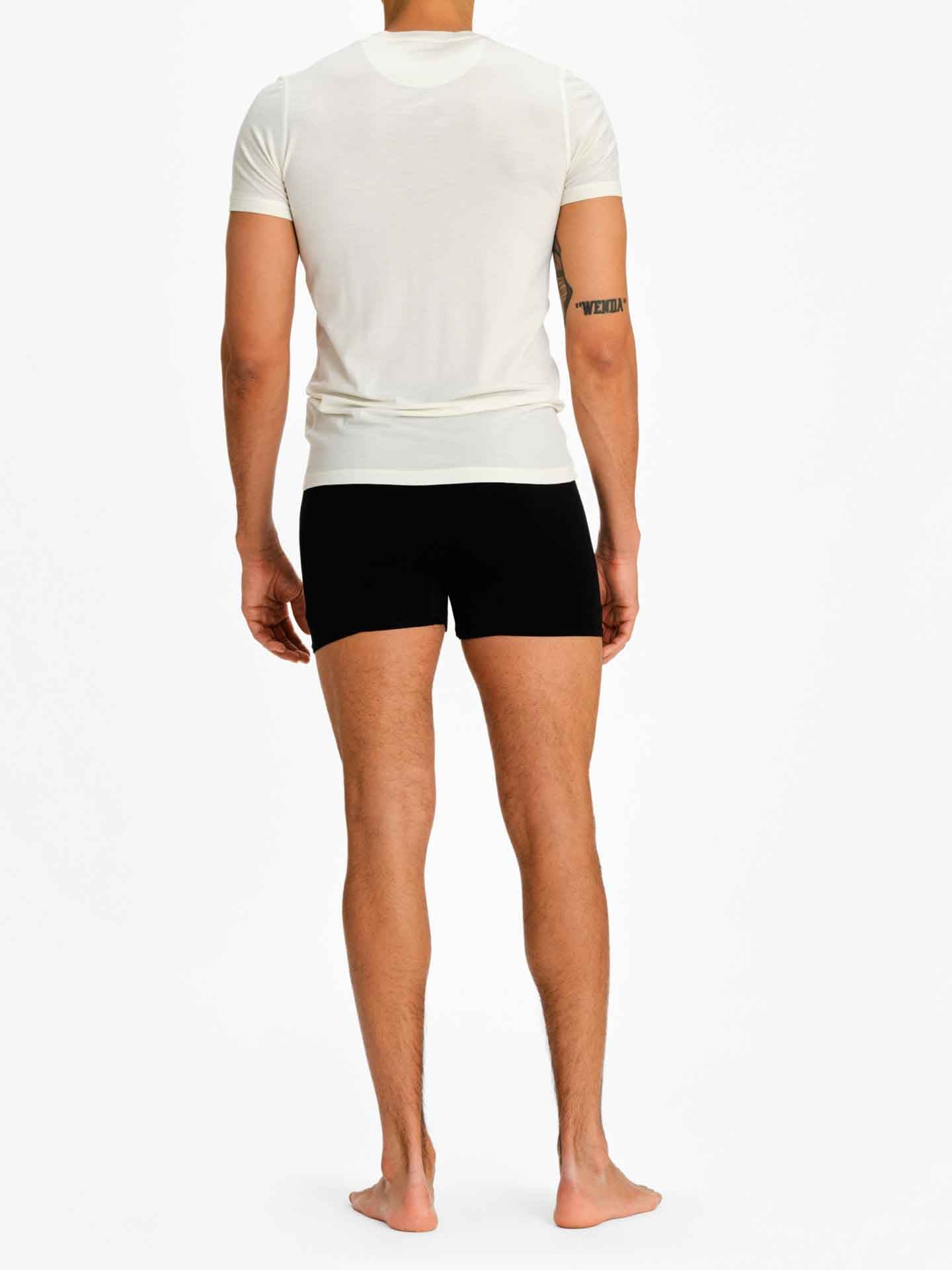 A person is standing barefoot against a plain white background, facing away from the camera. They are wearing a white Sno Merino Tee Men and black boxer briefs, part of our Scandinavian Luxury base-layers from We Norwegians. The person's left arm has a tattoo.