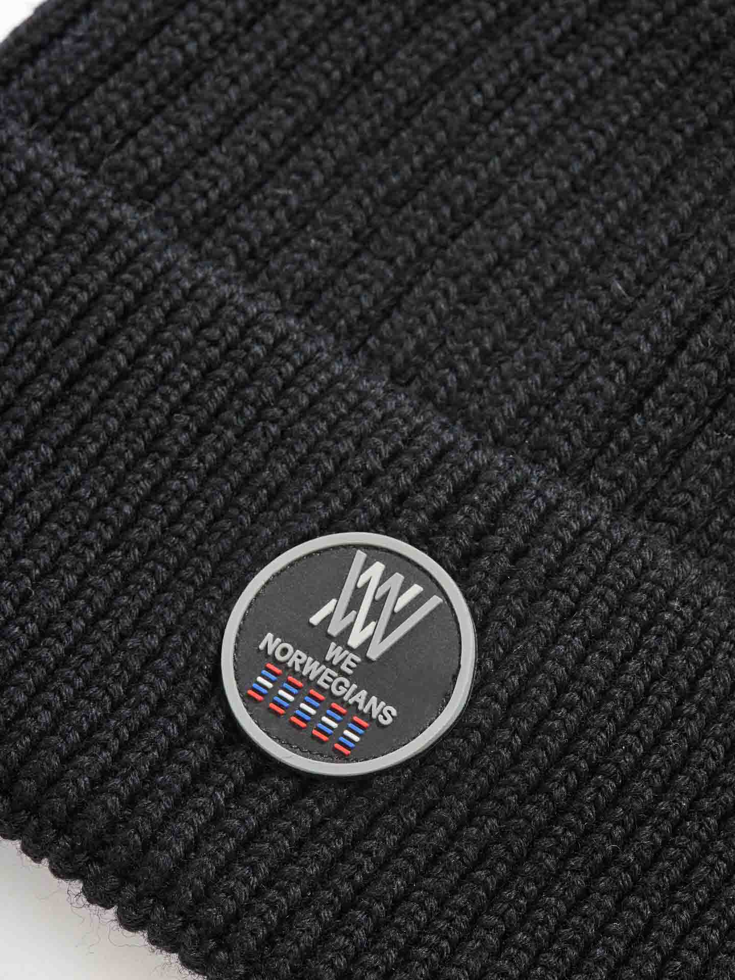 Close-up image of the Hemsedal Hat Women in black knit, showcasing a round black patch with white "XV" initials. Beneath, the text "WE NORWEGIANS" is displayed in white above a small design featuring red, white, and blue stripes. This hat is crafted by We Norwegians from premium merino wool for ultimate warmth and comfort.