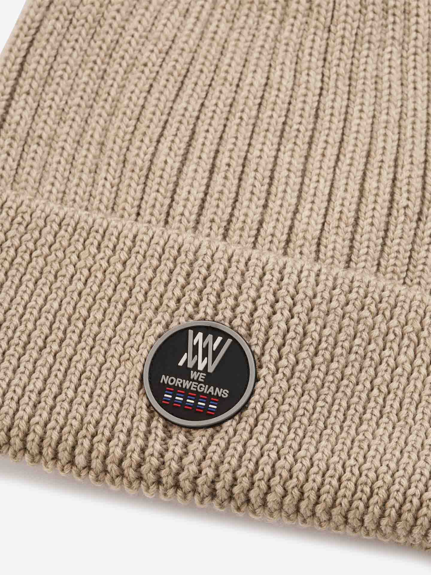 A close-up of the Hemsedal Hat for women by We Norwegians in beige, ribbed knit with a folded brim. The beanie features a round black patch displaying the "We Norwegians" brand name and logo, as well as a design of three stripes in red, white, and blue.