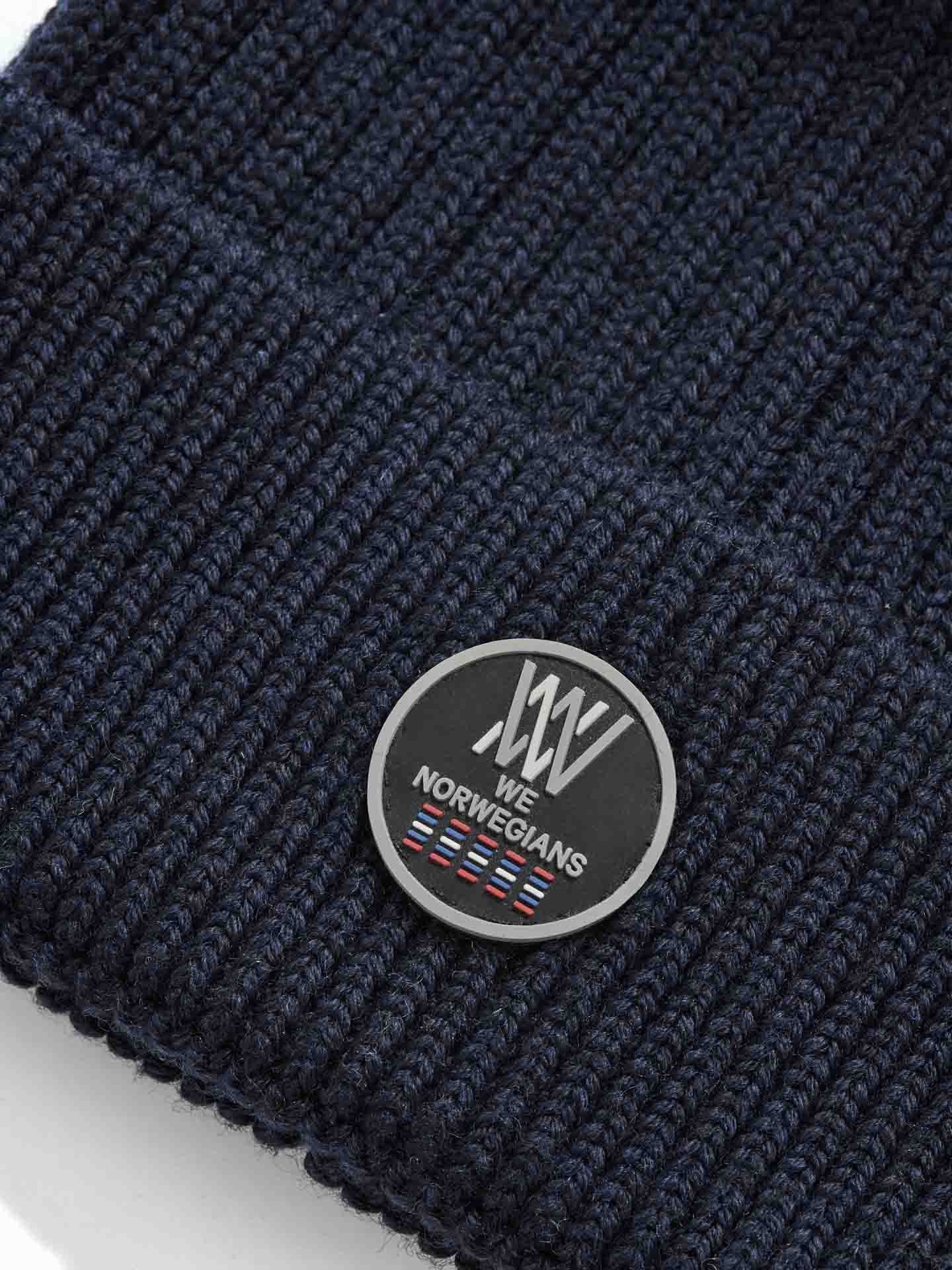 A close-up of the Hemsedal Hat Women by We Norwegians, in dark navy blue knit made from soft merino wool. The hat features a circular black patch with a white stylized logo, text reading "We Norwegians," and a small graphic of red, white, and blue stripes.