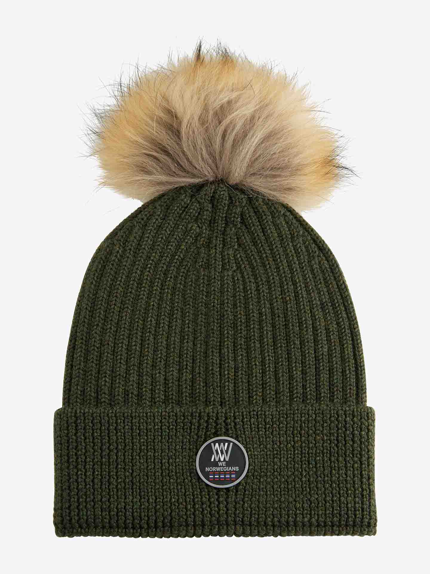 The Hemsedal Hat Women by We Norwegians is a green knitted winter hat crafted from cozy merino wool, topped with a large brown faux fur pom-pom. It features a folded cuff adorned with a round Norway-themed patch displaying a stylized design and text in the center, all set against a plain white background.