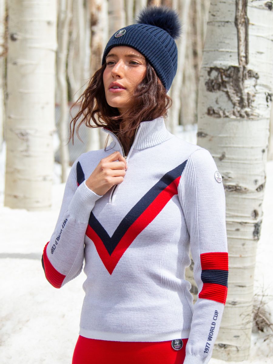 A person stands outdoors in a snowy forest, wearing a white sweater with red and black V-shaped stripes made from merino wool, red pants, and the We Norwegians' Hemsedal Hat Women in navy blue with a pom-pom. Birch trees are visible in the background. The person has their hand near the neck's zipper of the sweater.