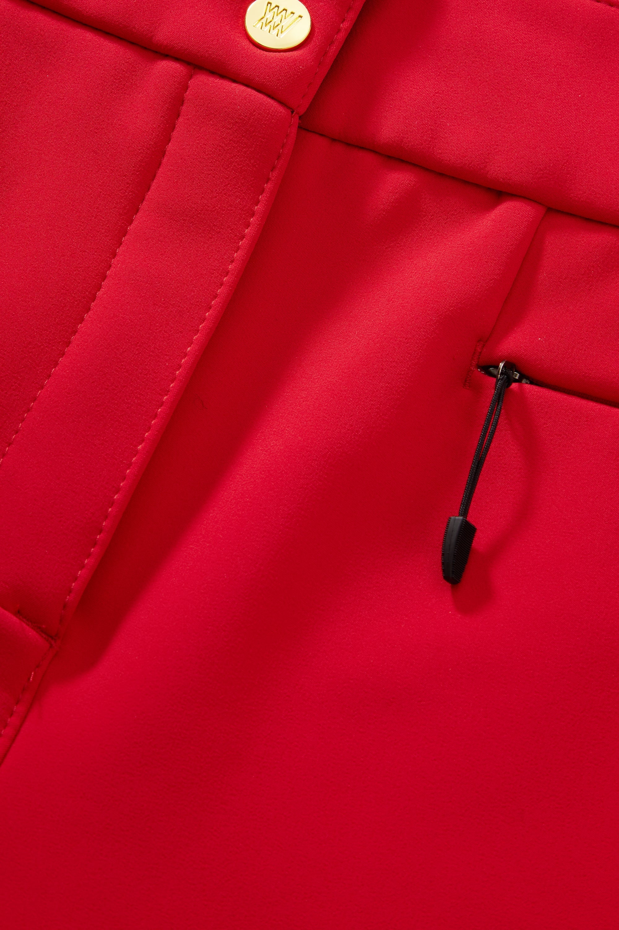 Close-up of a vibrant red garment, showcasing stitching details, a section of a pocket with a small drawstring cord and toggle, and a metallic button at the top. The fabric appears smooth and structured like the Kvitfjell Bootleg Ski Pant Women by We Norwegians.