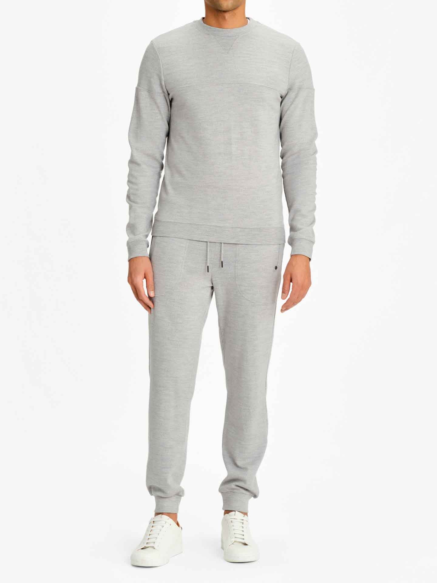 Person standing against a white background, wearing a light gray long-sleeved Tind Crewneck Men sweatshirt and matching Tind Joggers from We Norwegians, made from soft Merino wool. They are also wearing white sneakers. Their face is not visible in the image.