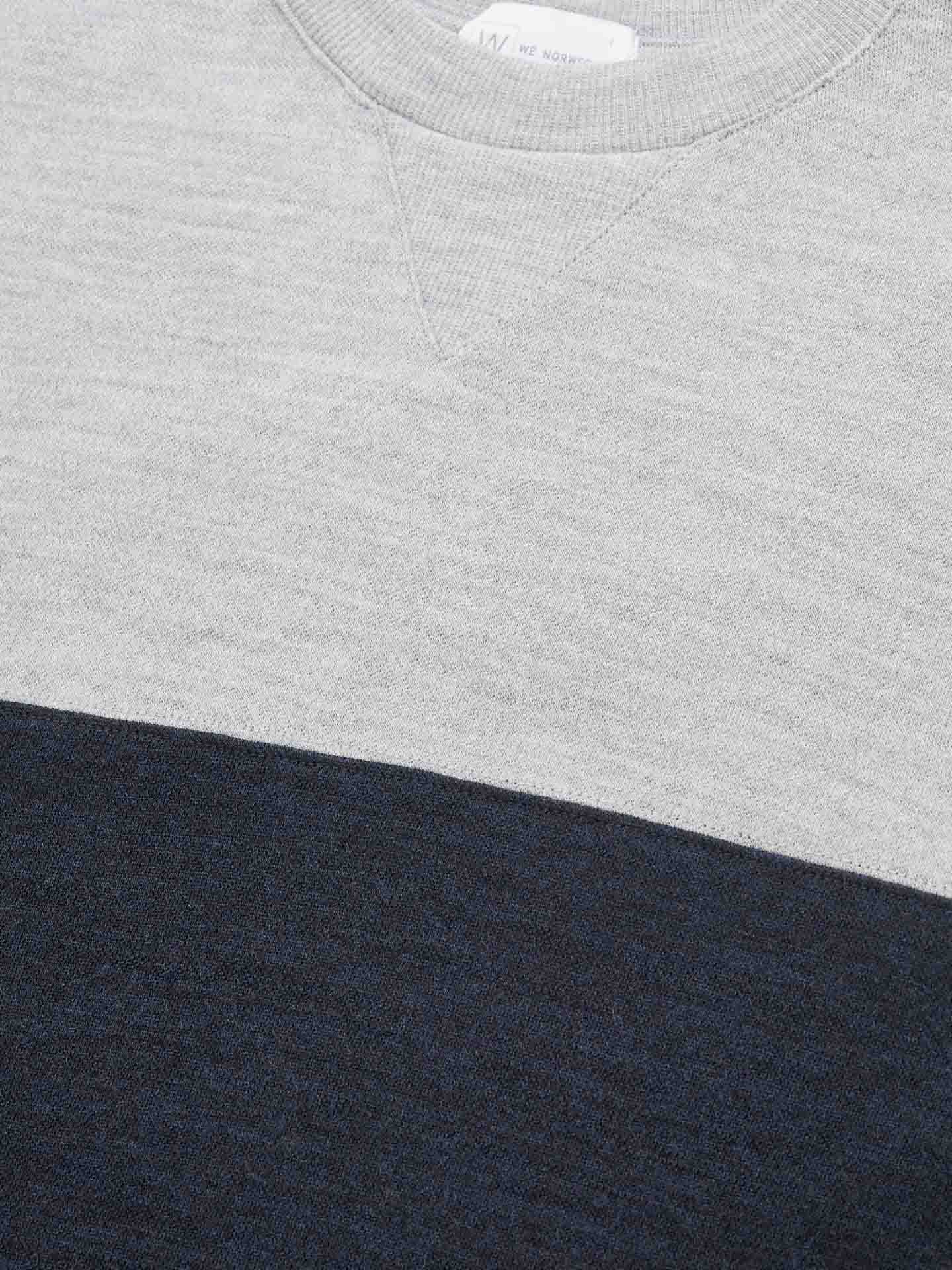 Close-up of the Tind Crewneck Men sweater by We Norwegians, showcasing a light grey upper section and a dark blue lower section made of Merino wool. The fabric texture is visible, and there's a small triangle detail at the neckline, making it perfect for pairing with Tind Joggers to complete your stylish sportswear look.