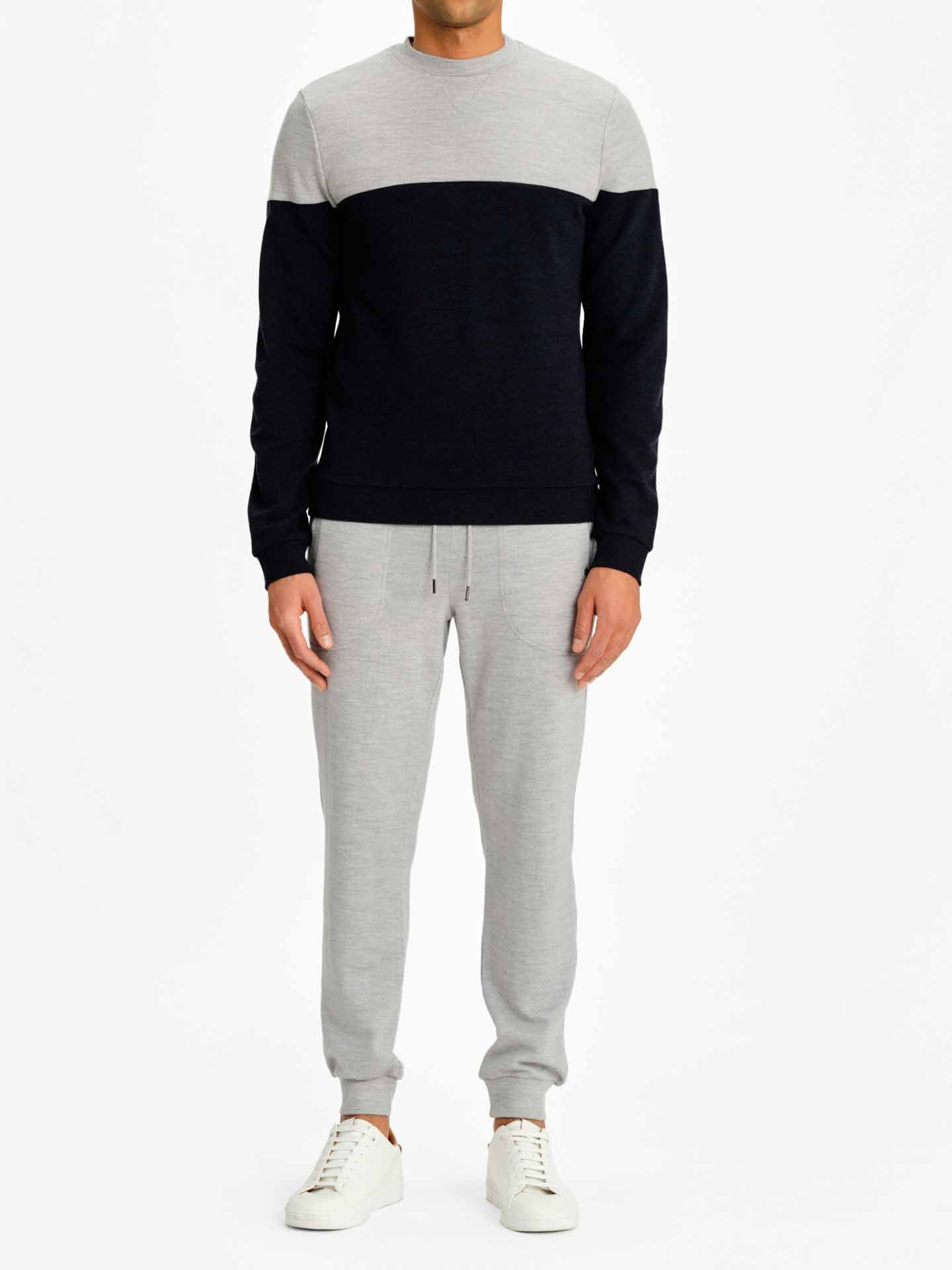 A person wearing a We Norwegians Tind Crewneck sweater, featuring a color-block design with the upper part in light gray and the lower part in dark navy blue, paired with Tind Joggers and white sneakers. The person is standing against a plain white background.