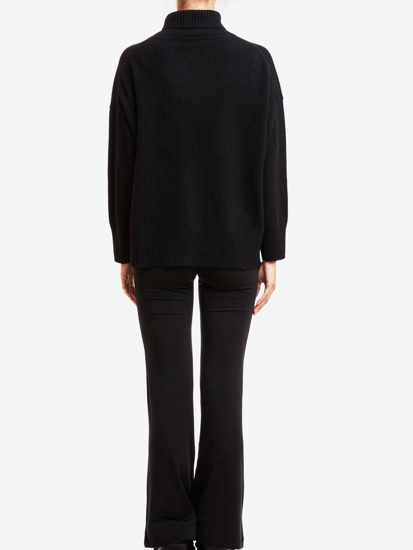 A person is standing with their back to the camera, wearing a We Norwegians Blefjell Sweater Women in black paired with fitted black pants. The background is plain white.