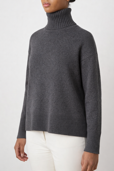 A person wears the We Norwegians Blefjell Sweater Women—Pure Cashmere in dark gray, featuring ribbed cuffs and hem, paired with white pants against a light background for an effortless luxury look.